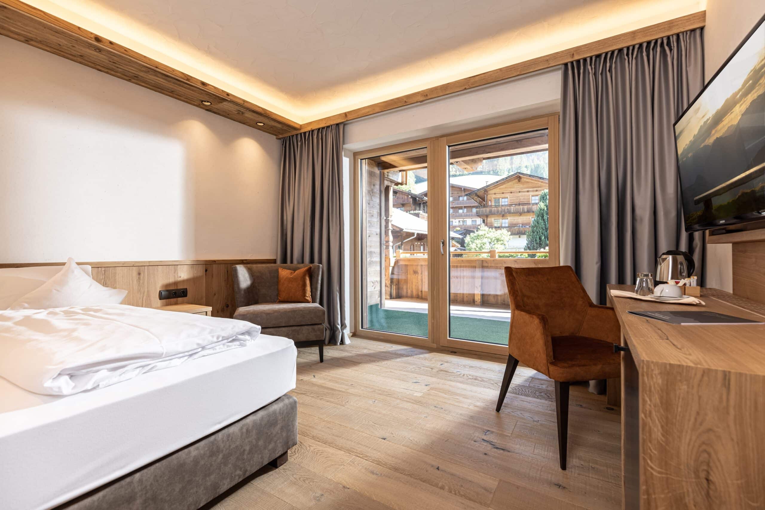 Ein modernes Hotelzimmer mit einem großen Bett, hölzernen Möbeln und bodentiefen Fenstern, die auf einen Balkon mit Blick auf alpine Chalets führen.