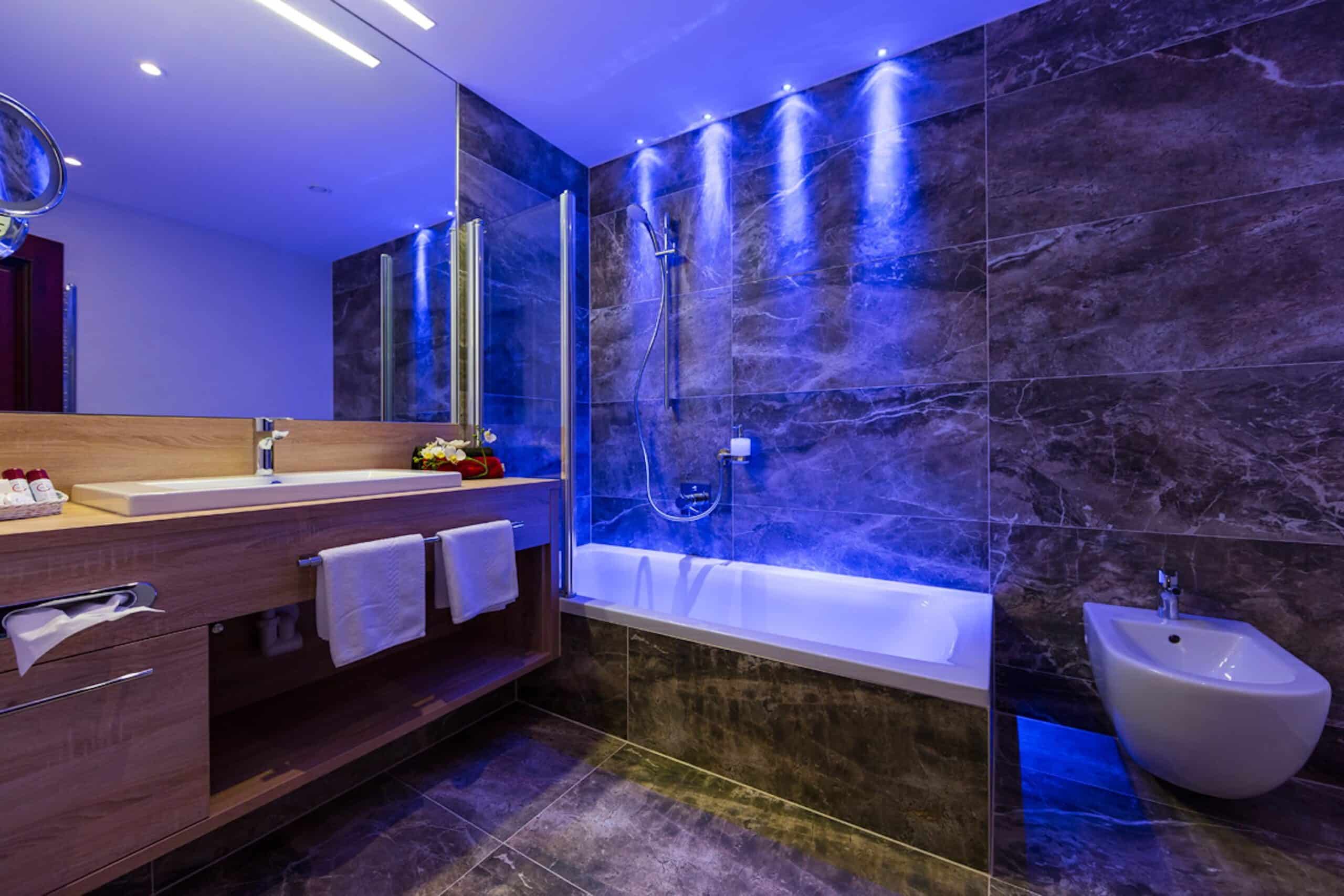 Modernes Badezimmer mit dunklen Marmorwänden, beleuchtet mit blauen LEDs, ausgestattet mit einer Badewanne, einem Waschbecken mit Unterschrank und einem Wand-WC.