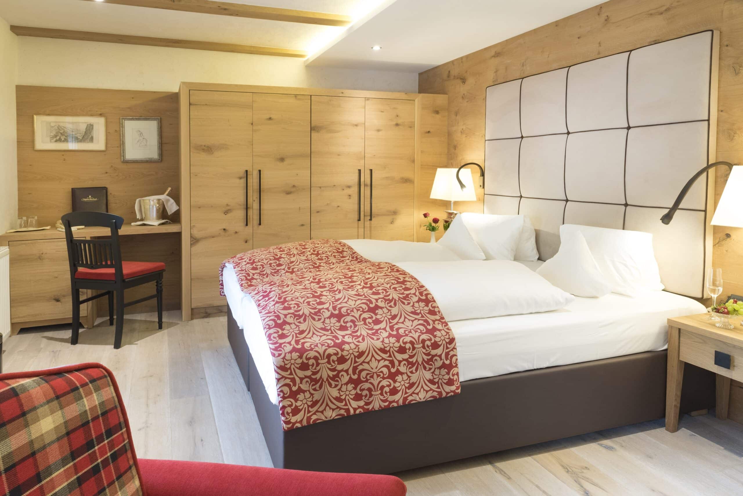 Gemütliches Hotelzimmer mit einem großen Doppelbett mit rot gemustertem Überwurf, holzvertäfelten Wänden und modernen Möbeln.