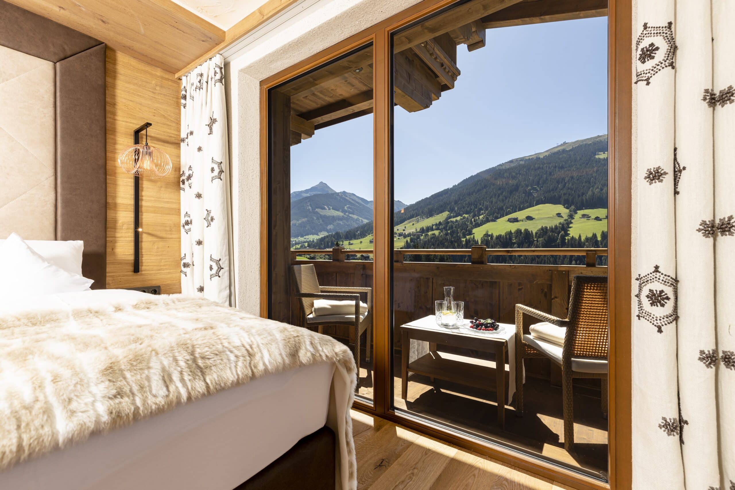 Blick aus einem gemütlichen Hotelzimmer mit aufgeschlagener Balkontür, die zu einem Balkon mit Sitzgelegenheiten führt. Draußen sieht man eine malerische Berglandschaft an einem sonnigen Tag.