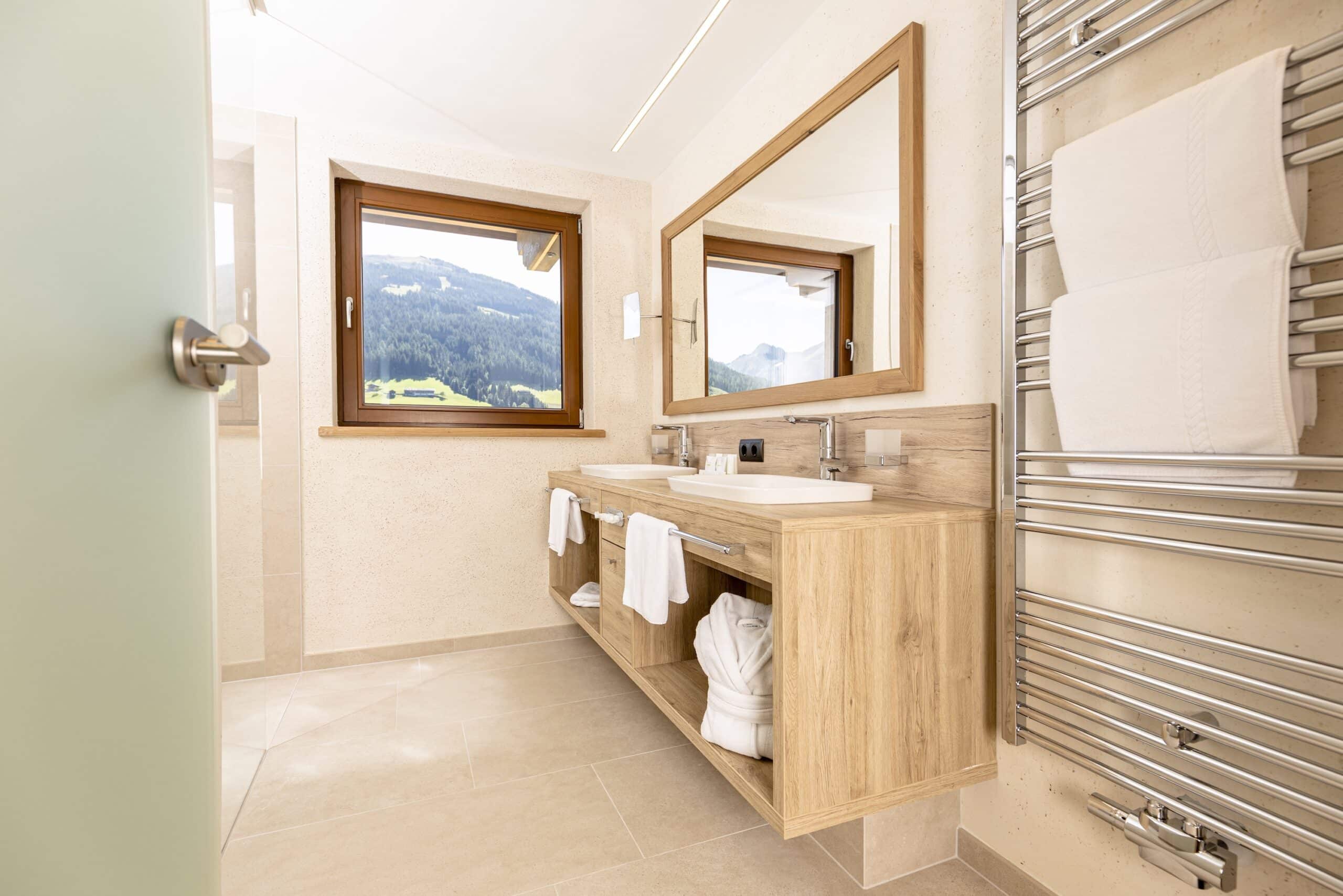 Helles, modernes Badezimmer mit Holzschränken, zwei Spiegeln und einem Blick auf die Berge durch das Fenster.