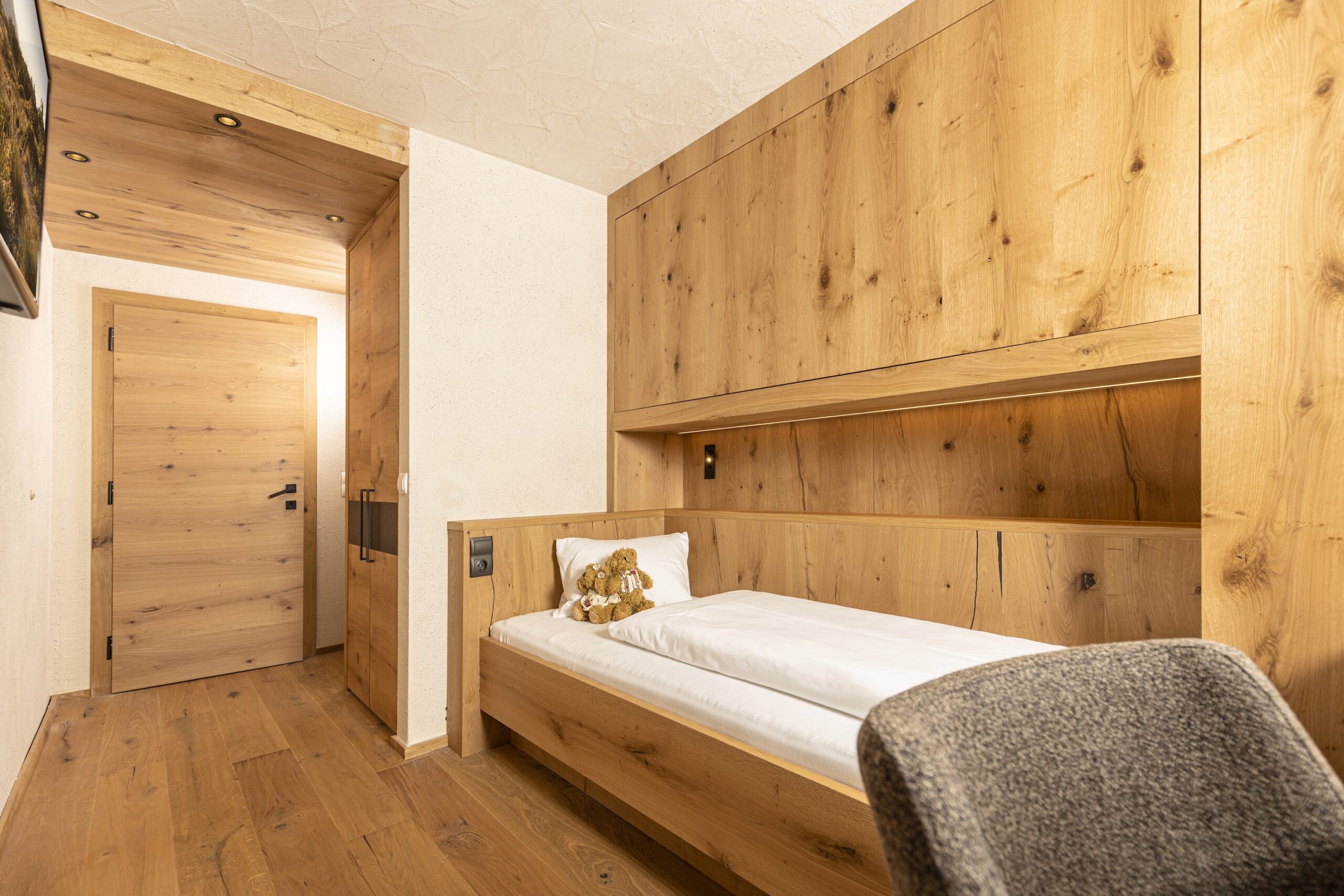 Gemütliches Schlafzimmer mit Holzwänden und einem Einzelbett, daneben ein grauer Stuhl.