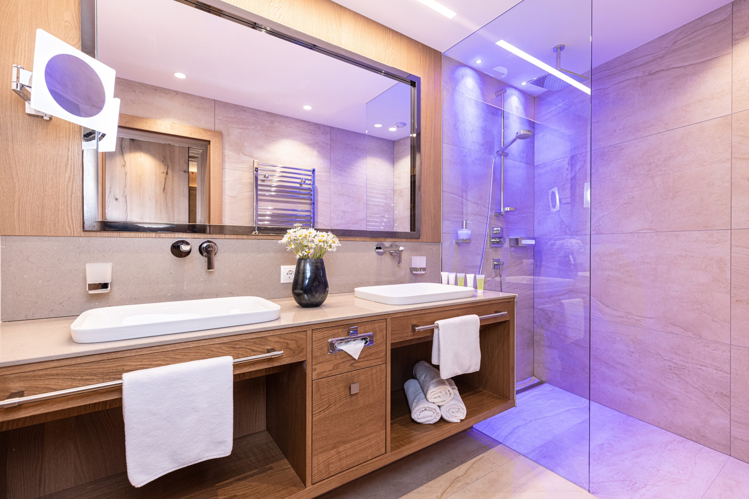 Modernes Badezimmer mit Doppelwaschbecken, Holzunterschrank, großen Spiegeln und einer beleuchteten Duschkabine.