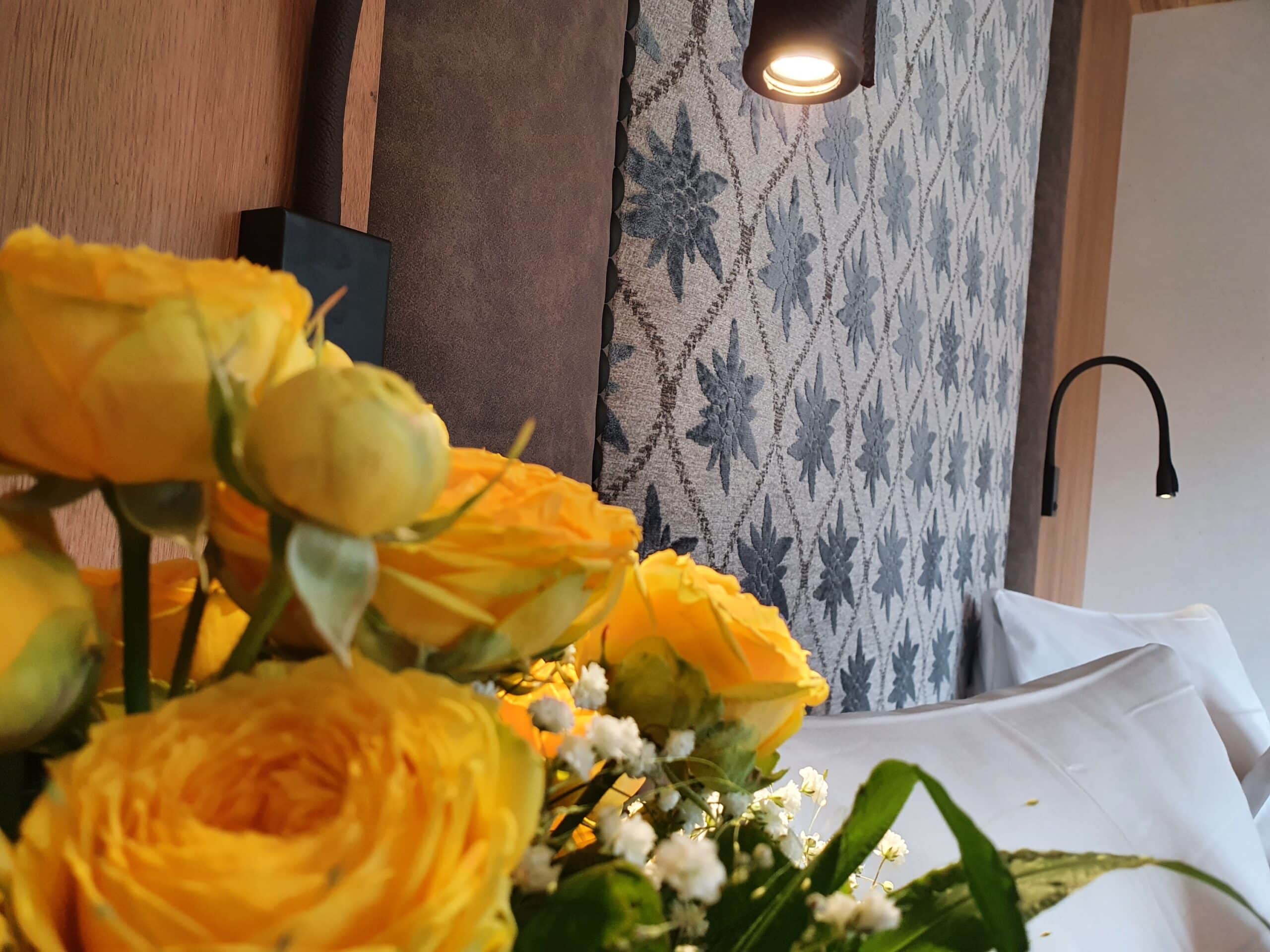 Ein Arrangement von leuchtend gelben Rosen im Vordergrund, dahinter eine gemütliche Ecke eines Zimmers mit dekorativer Tapete und moderner Beleuchtung.