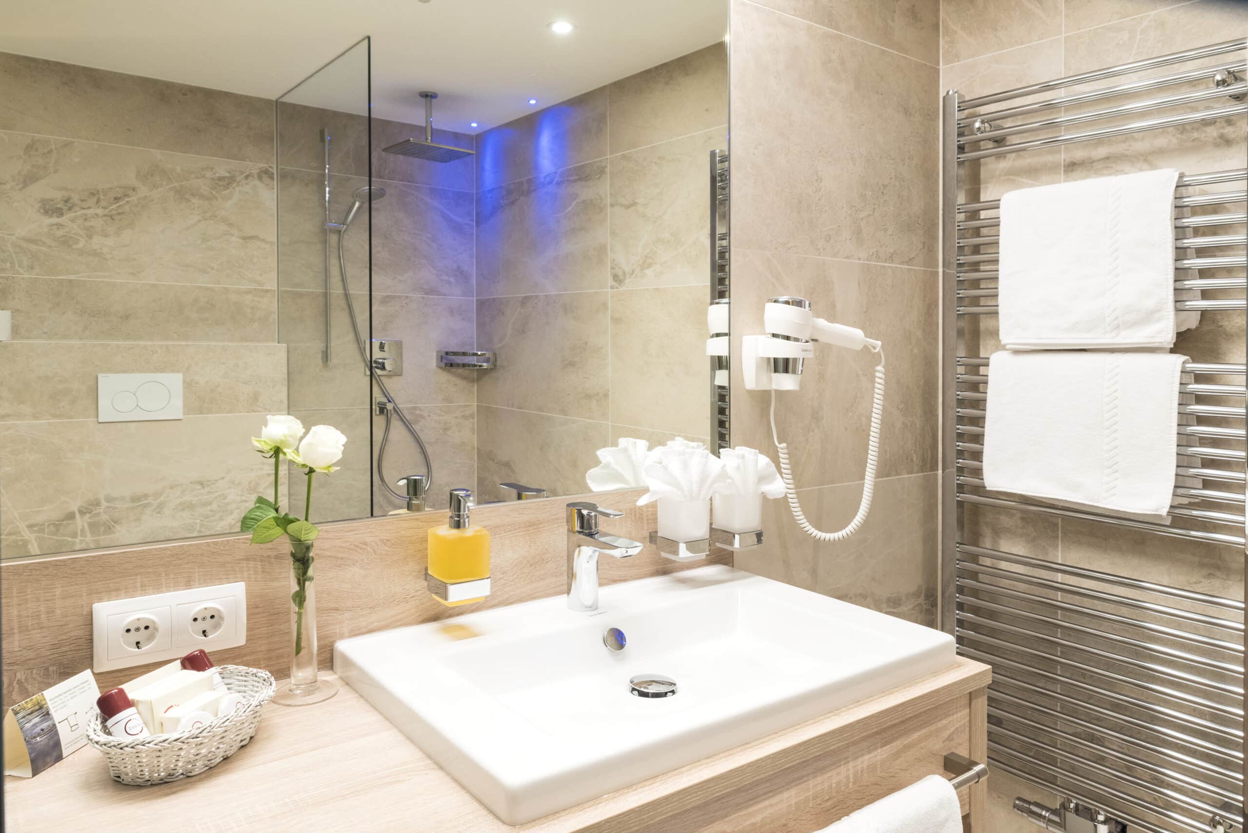 Modernes Badezimmer mit großer begehbarer Dusche, beleuchtet in sanftem Blau, sowie einem weißen Waschbecken, Handtüchern und diversen Badutensilien.