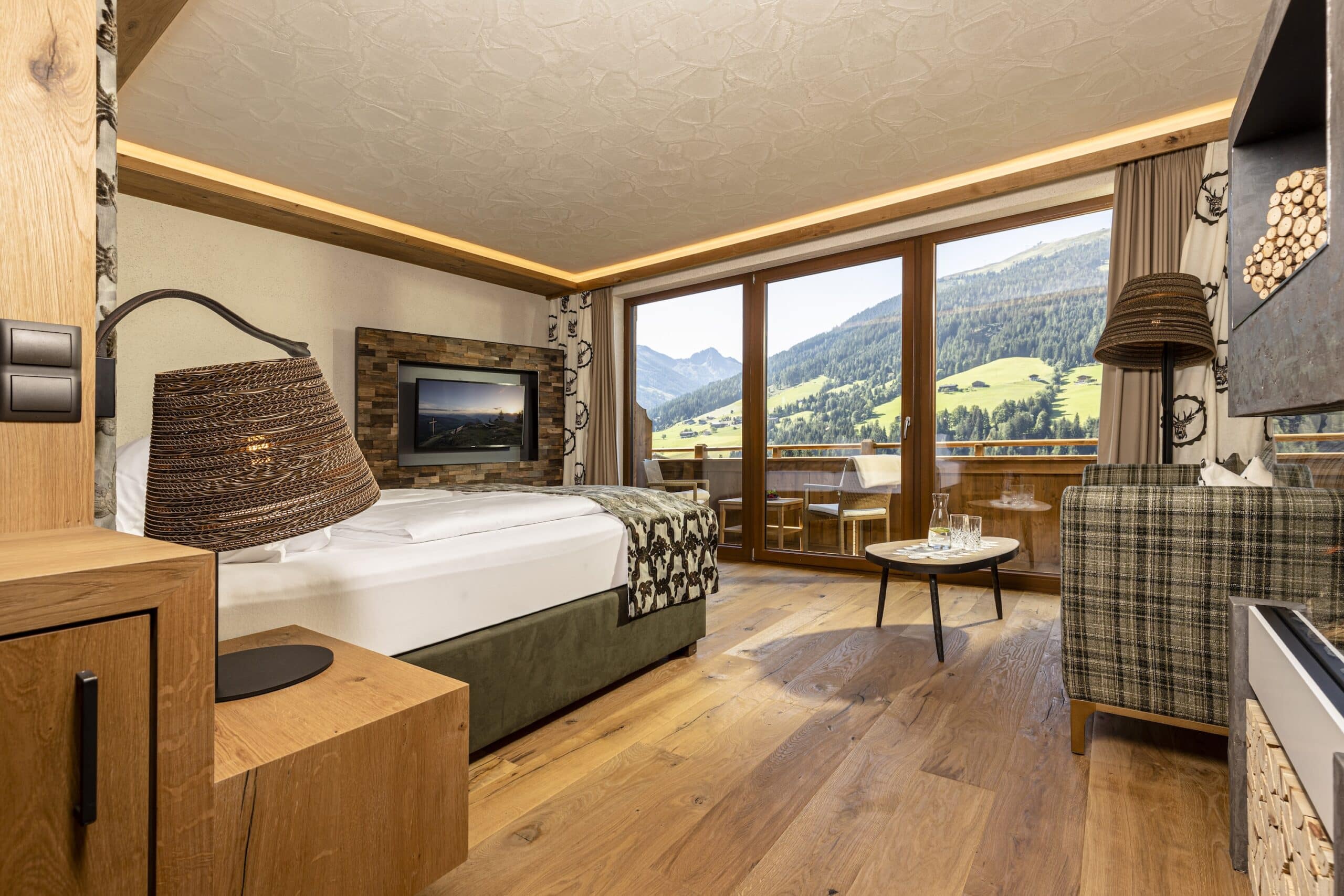 Ein modern eingerichtetes Hotelzimmer mit einem großen Bett, Holzboden und Panoramafenstern, die einen Blick auf eine malerische Berglandschaft bieten. Ein gemütlicher Sitzbereich ist ebenfalls zu sehen.