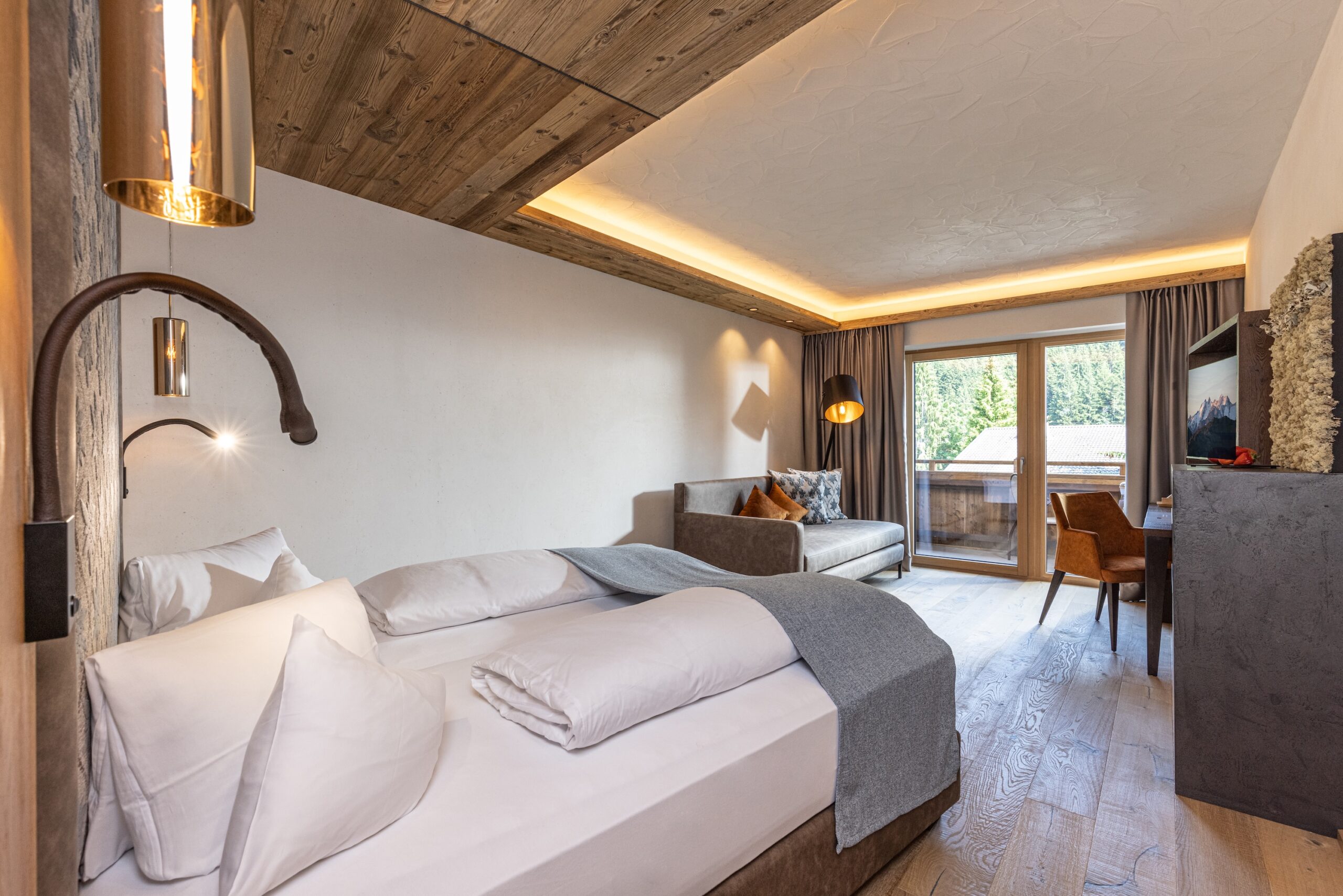 Ein gemütliches Schlafzimmer in einem modernen Stil mit einem großen Doppelbett, grauer Bettwäsche, hölzernen Decken und einem Arbeitsbereich mit Fensterblick.