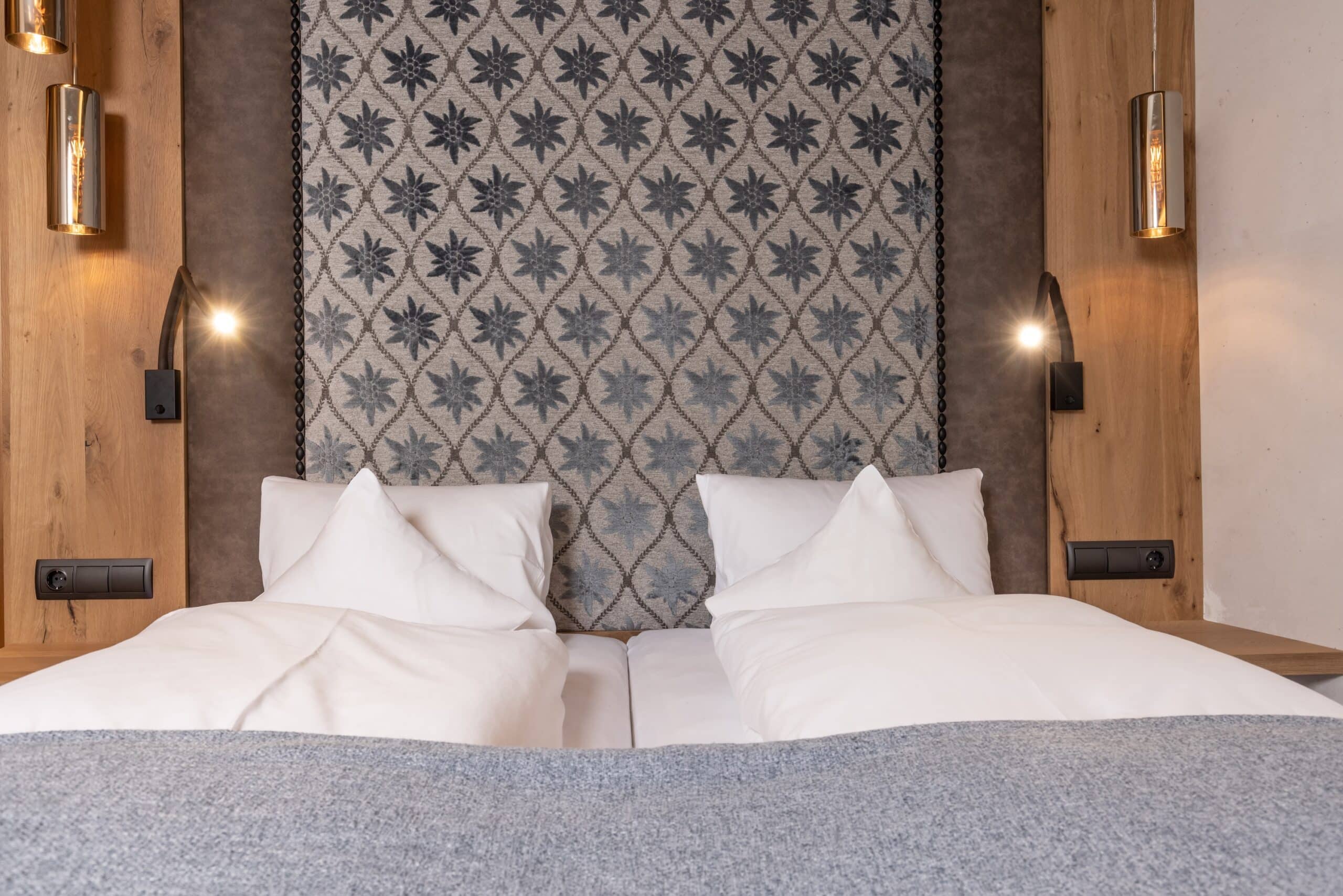 Ein gemütliches Hotelzimmer mit einem großen Doppelbett, weißen Kissen und einer grauen Bettdecke. Hinter dem Bett hängt ein dekorativer Wandteppich mit Sternenmustern. Beidseitig sind moderne Wandlampen angebracht.