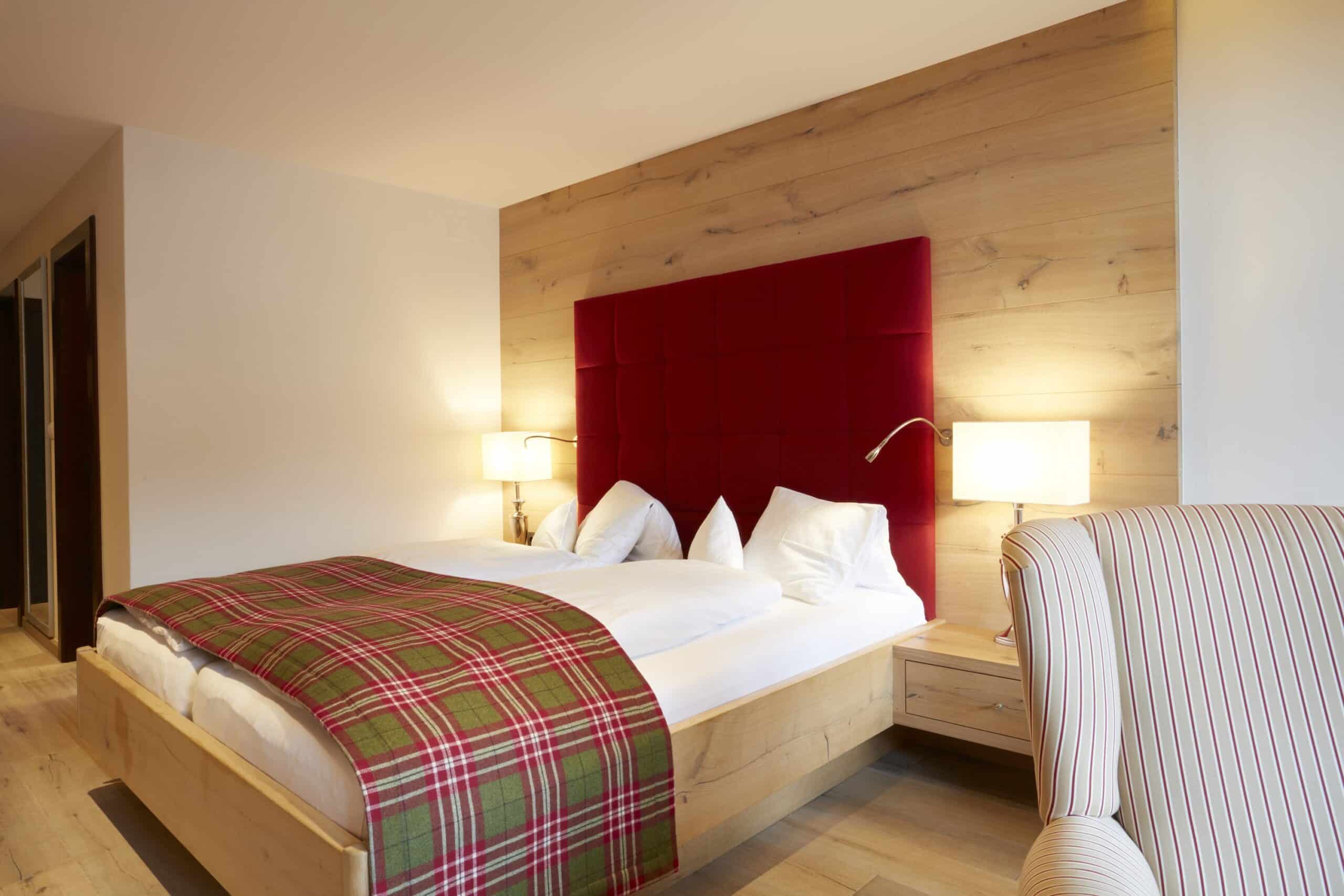 Modernes Hotelzimmer mit einem großen Holzbett mit rotem Kopfteil, weißer Bettwäsche und einem karierten Überwurf, neben einem gestreiften Sessel.