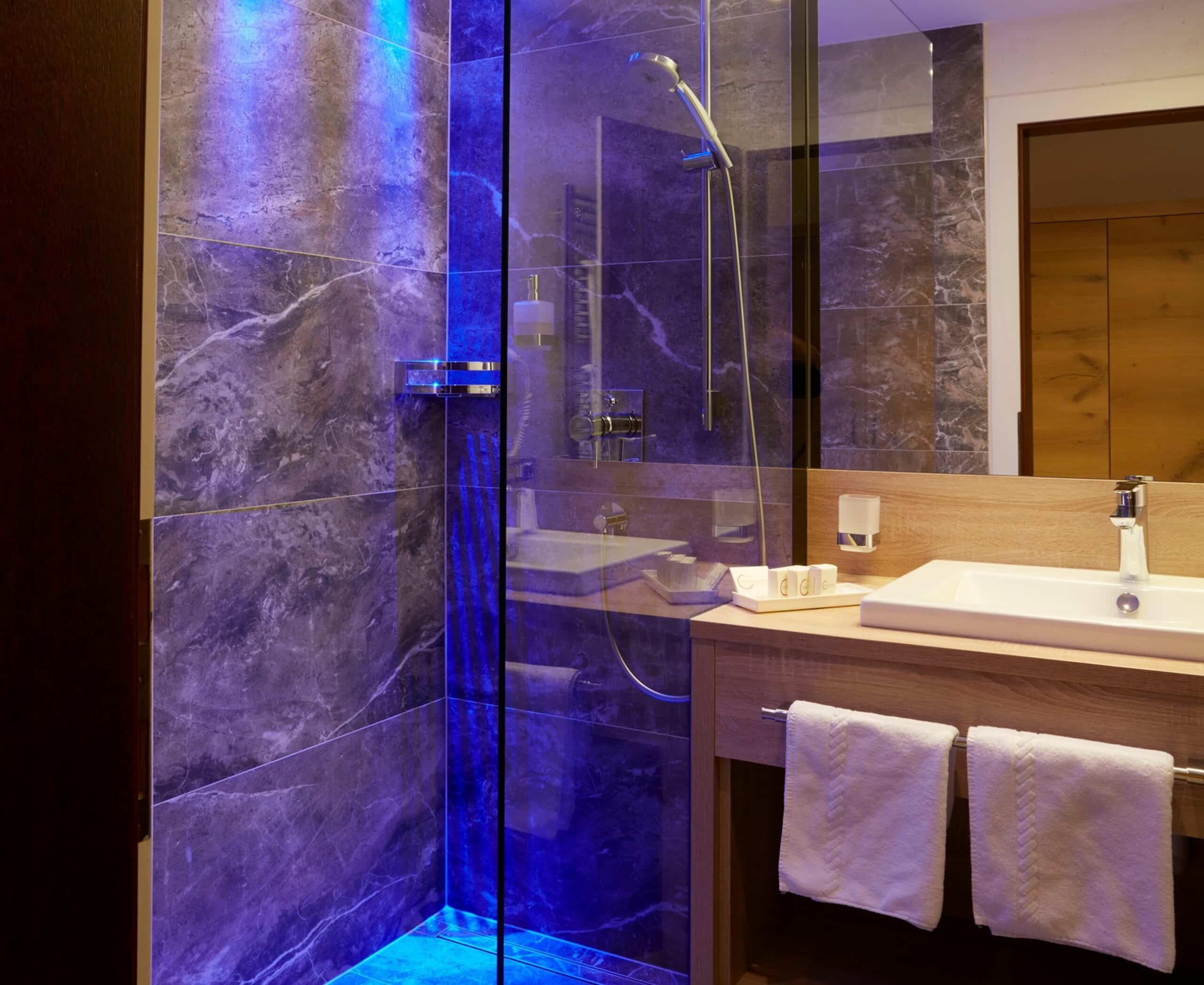 Modernes Badezimmer mit beleuchteter Duschkabine aus Glas, dunklen Marmorfliesen und einem Waschbecken mit Spiegel.