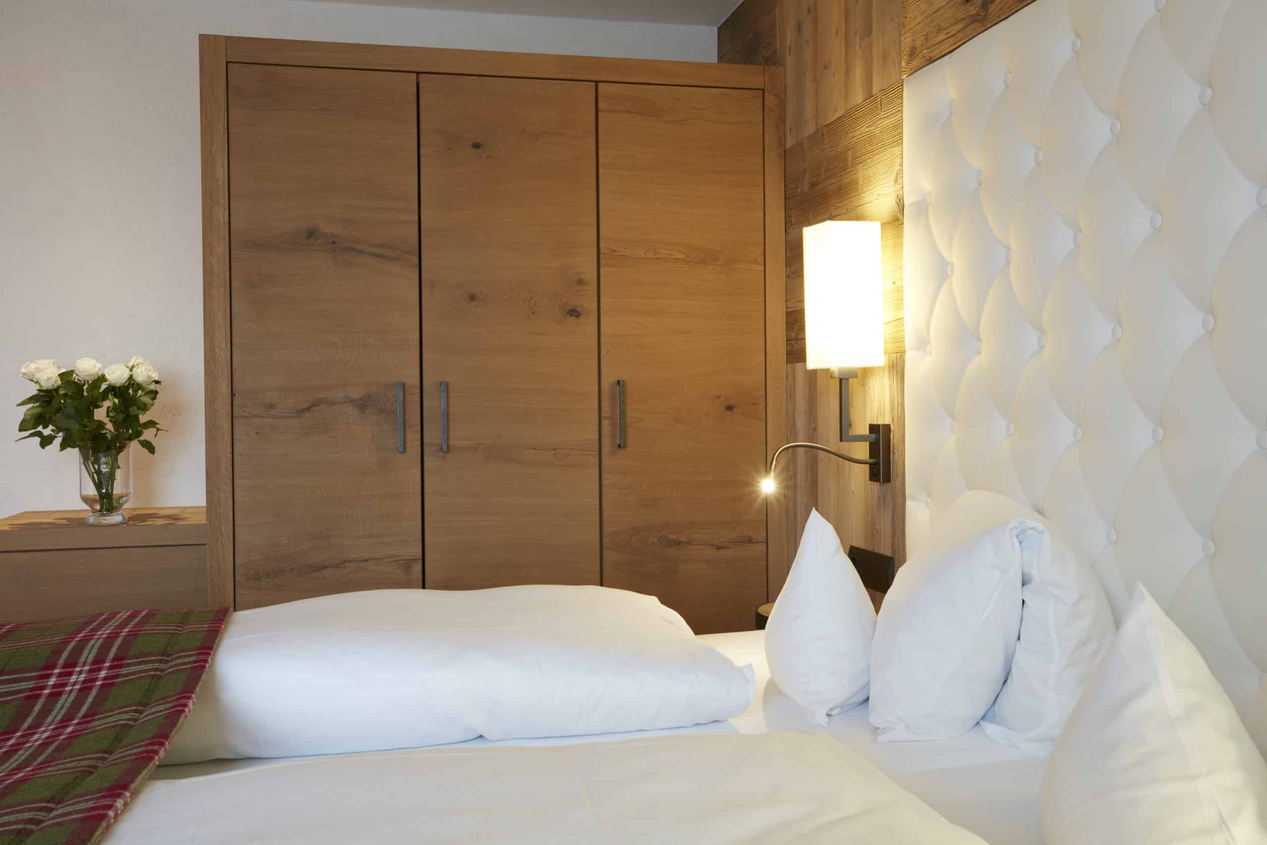 Ein gemütlich eingerichtetes Schlafzimmer mit einem großen Doppelbett, weißen Kissen und einer hölzernen Schrankwand. Ein Nachttisch mit einer leuchtenden Lampe und frischen Blumen befindet sich neben dem Bett.