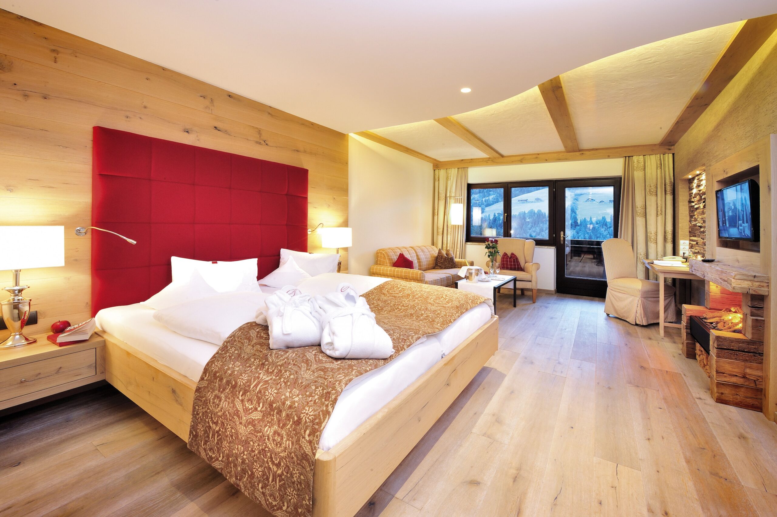 Luxuriöses Hotelzimmer mit großem Doppelbett, rotem Kopfteil und Holzboden. Panoramafenster bieten Ausblick auf eine Berglandschaft.