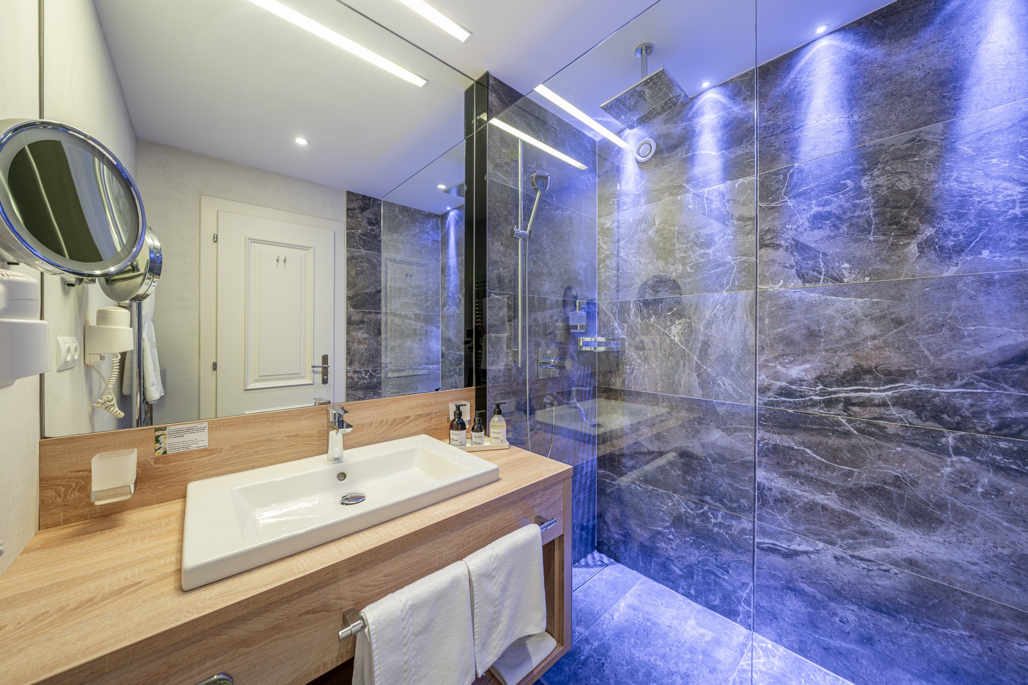 Badezimmer mit ebenerdiger Dusche in blauem Licht und Single-Waschbecken auf hölzernem Waschtisch.