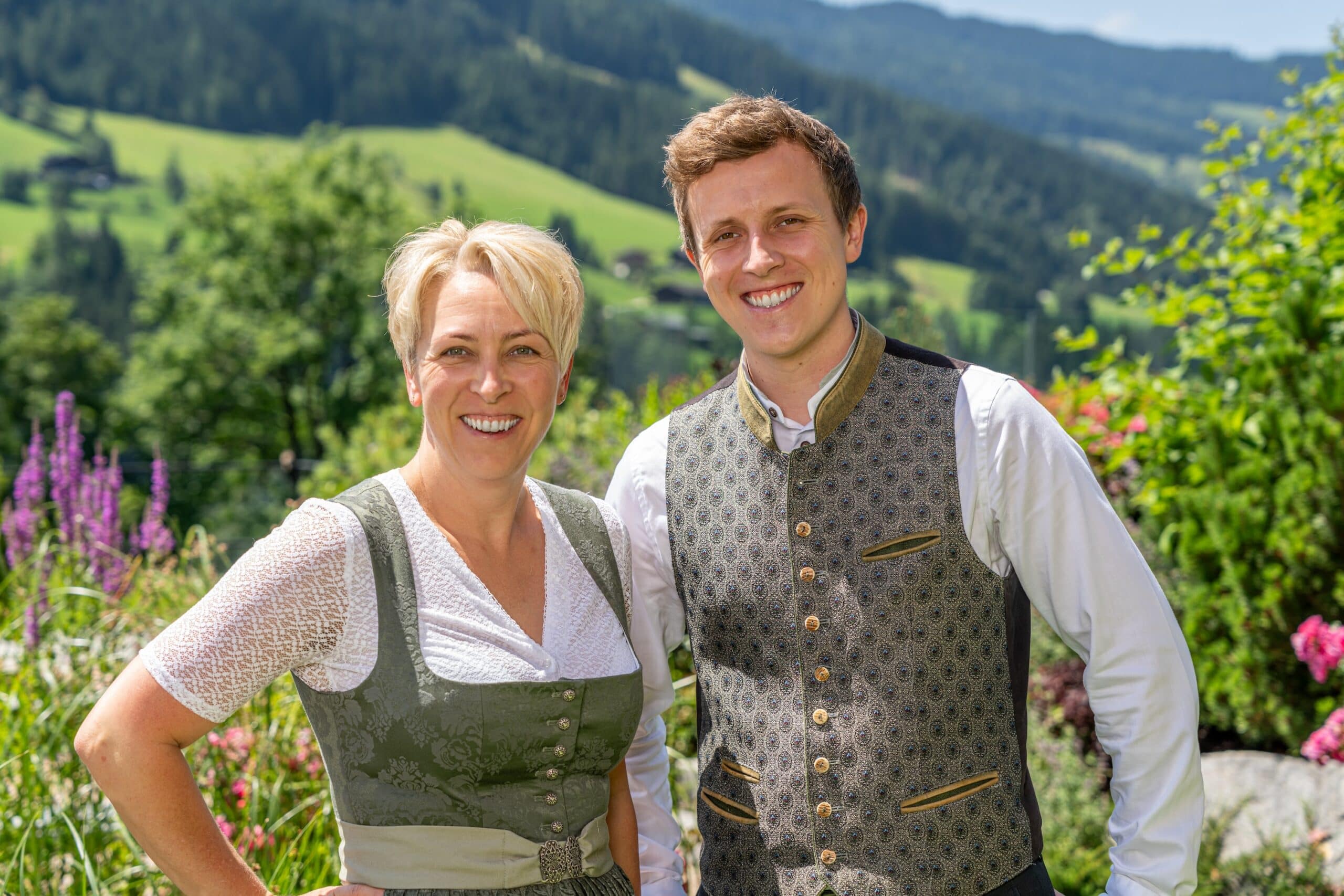 Ein älterer Mann und eine jüngere Frau in traditioneller Tracht stehen lächelnd vor einer malerischen Berglandschaft.