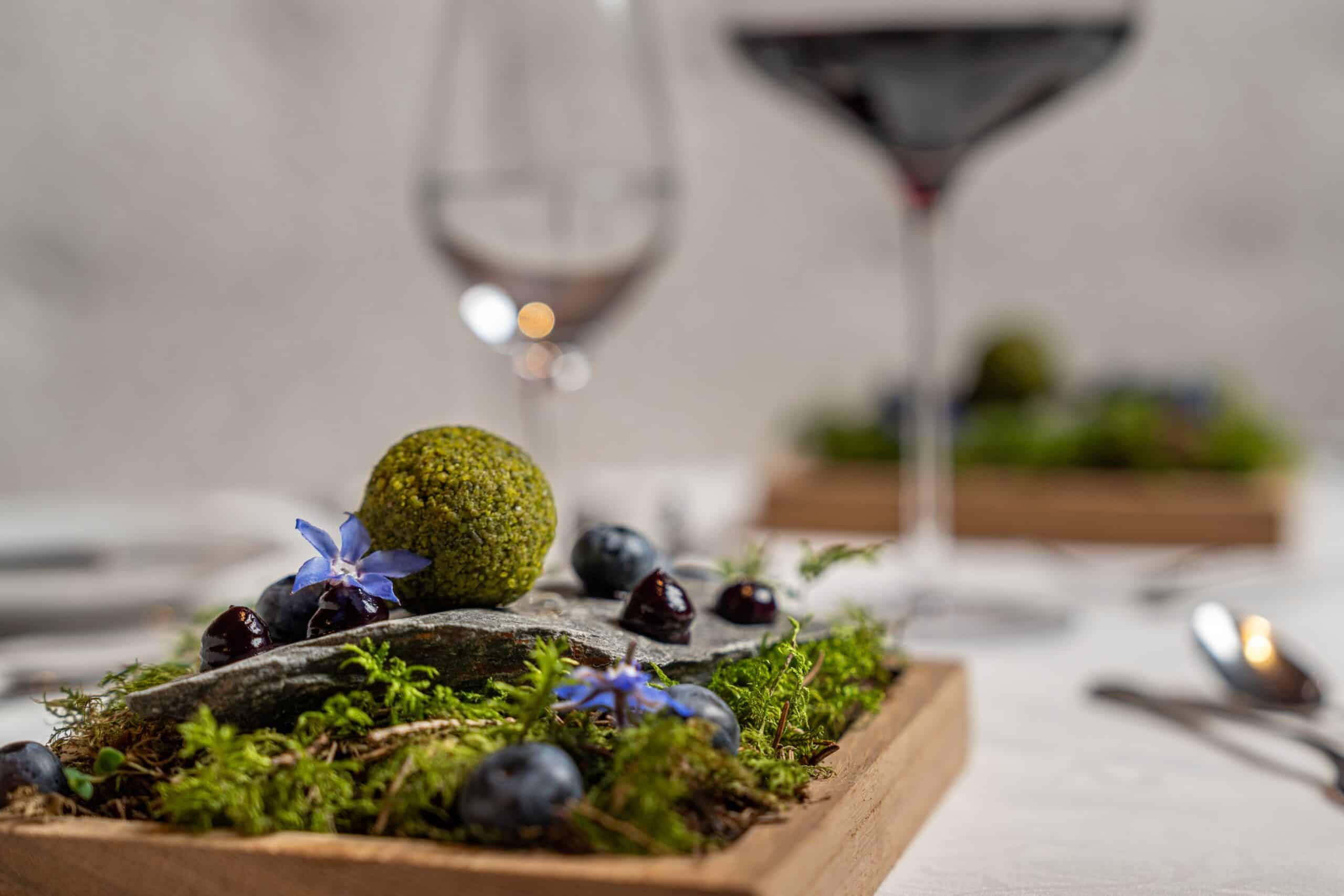 Ein Gourmet-Dessert mit Heidelbeeren und essbaren Blumen, angerichtet auf einem hölzernen Tablett, im Hintergrund unscharf sichtbare Weingläser.