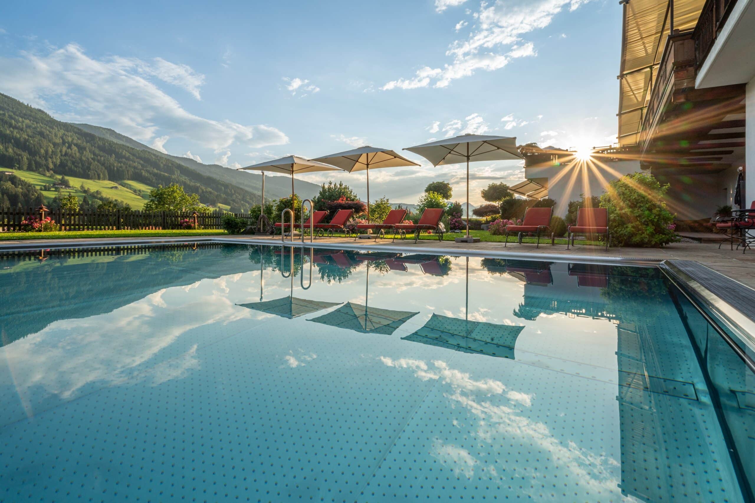 Ein Außenschwimmbad mit klarem Wasser, umgeben von Sonnenliegen und Sonnenschirmen, mit Blick auf grüne Hügel und einem spektakulären Sonnenuntergang im Hintergrund.