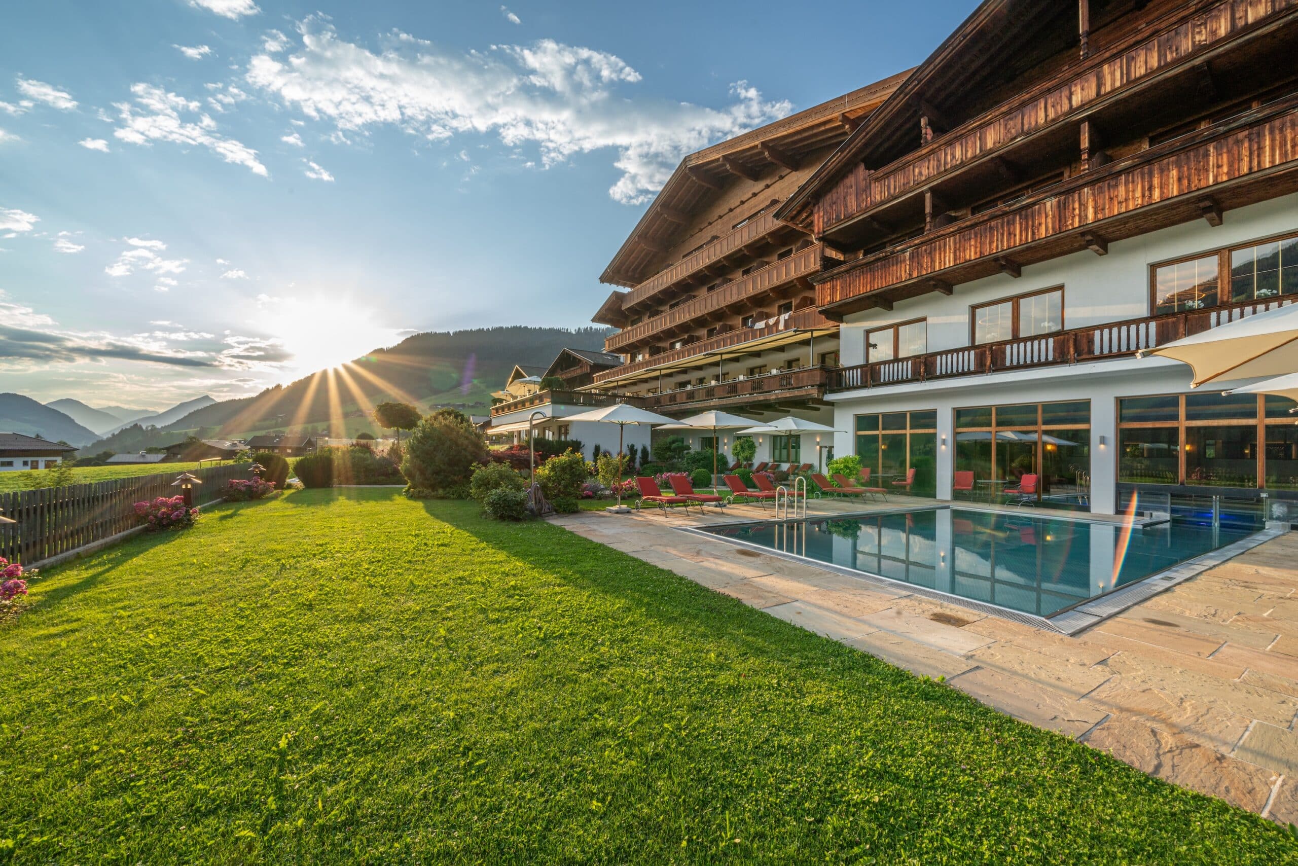 Sonnenuntergang hinter einem großen Hotel mit Holzfassade, vorne ein Swimmingpool und eine gepflegte Rasenfläche, umgeben von Bergen.