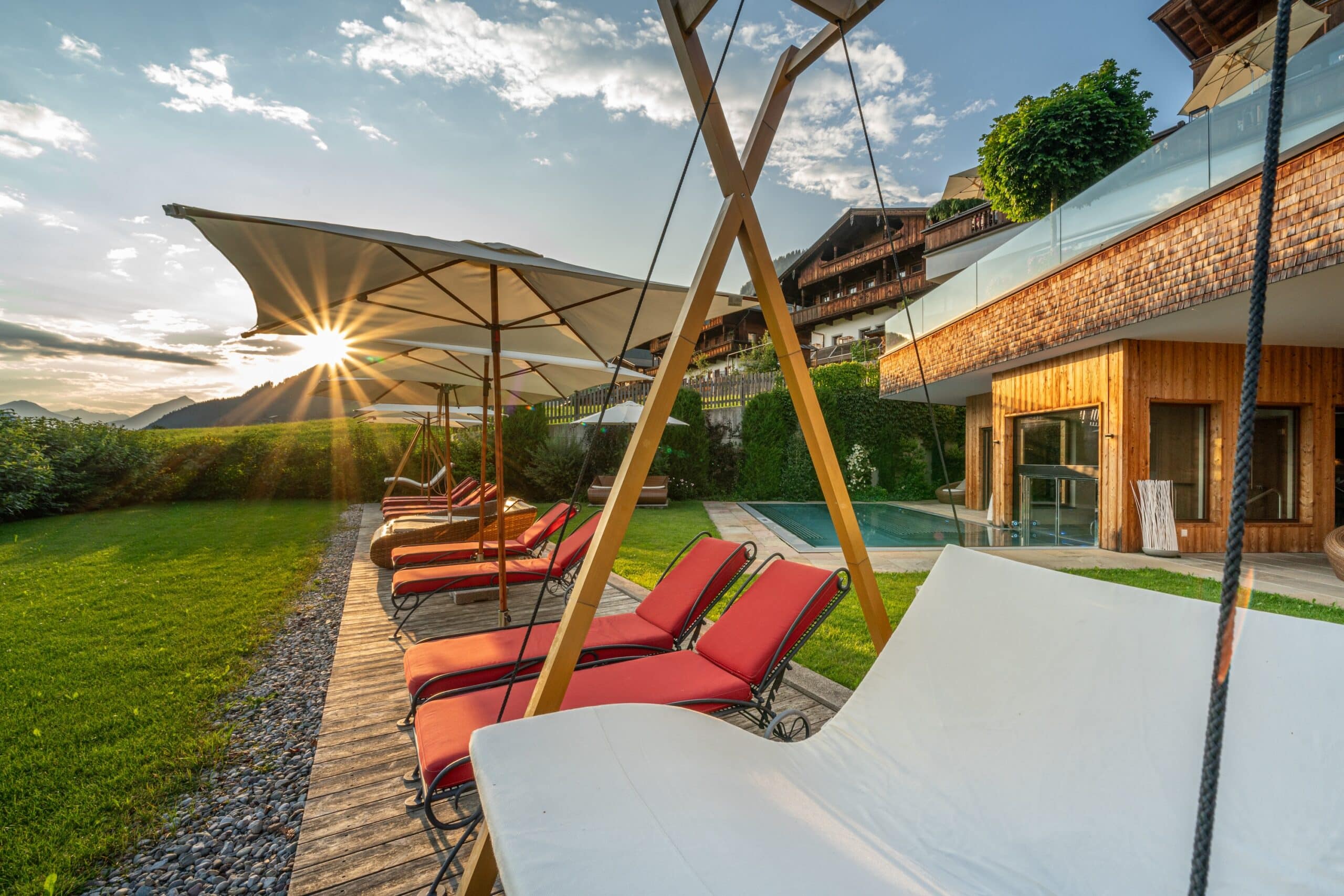 Sonnenaufgang über einem luxuriösen Garten mit Liegestühlen und Sonnenschirmen vor einem modernen Haus mit Holzfassade und grünen Hügeln im Hintergrund.