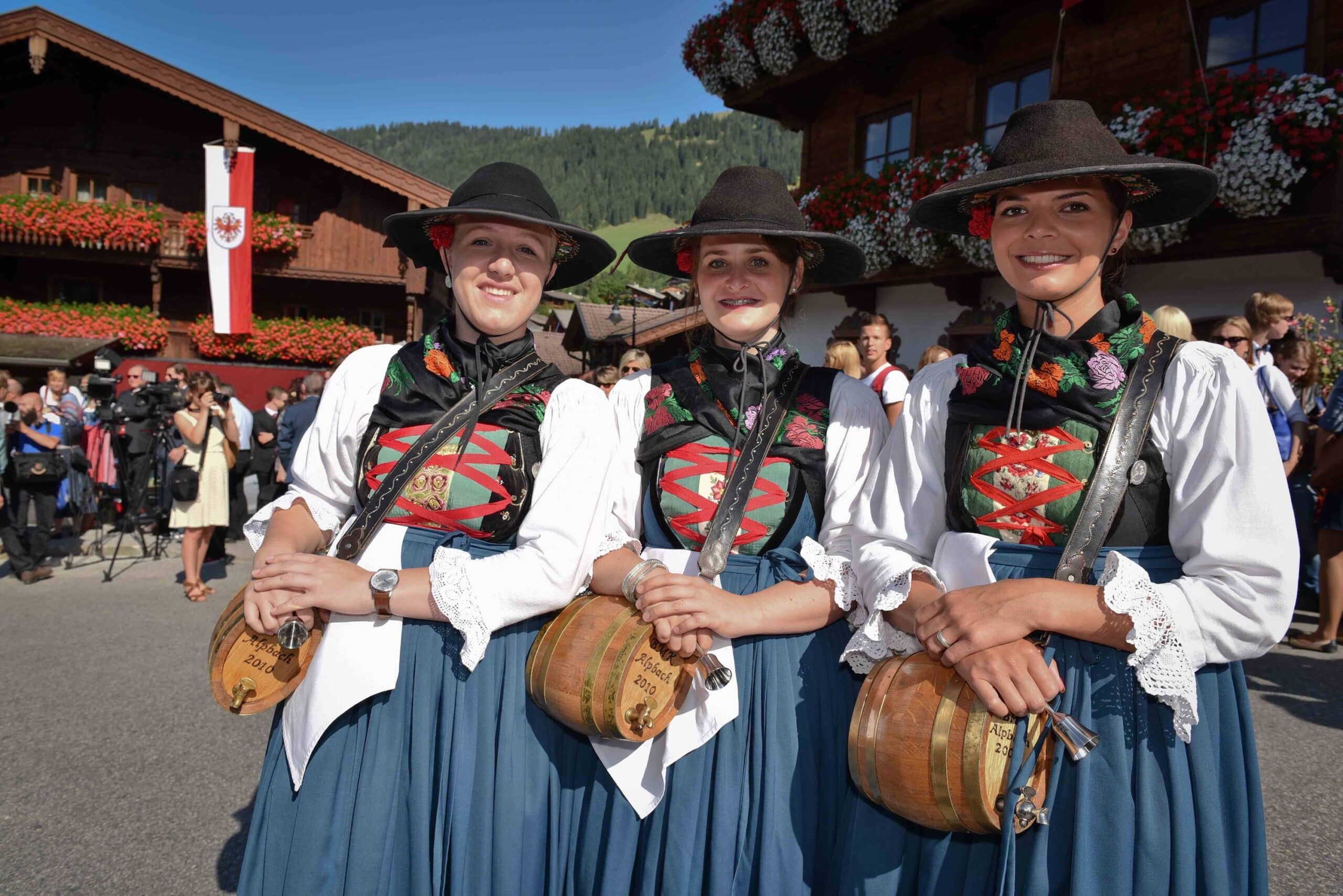 Drei Frauen in traditioneller bayerischer Tracht mit Hüten, die bei einem Volksfest lächeln. Sie tragen jeweils eine Kuhglocke und stehen vor einem blumenverzierten Haus.