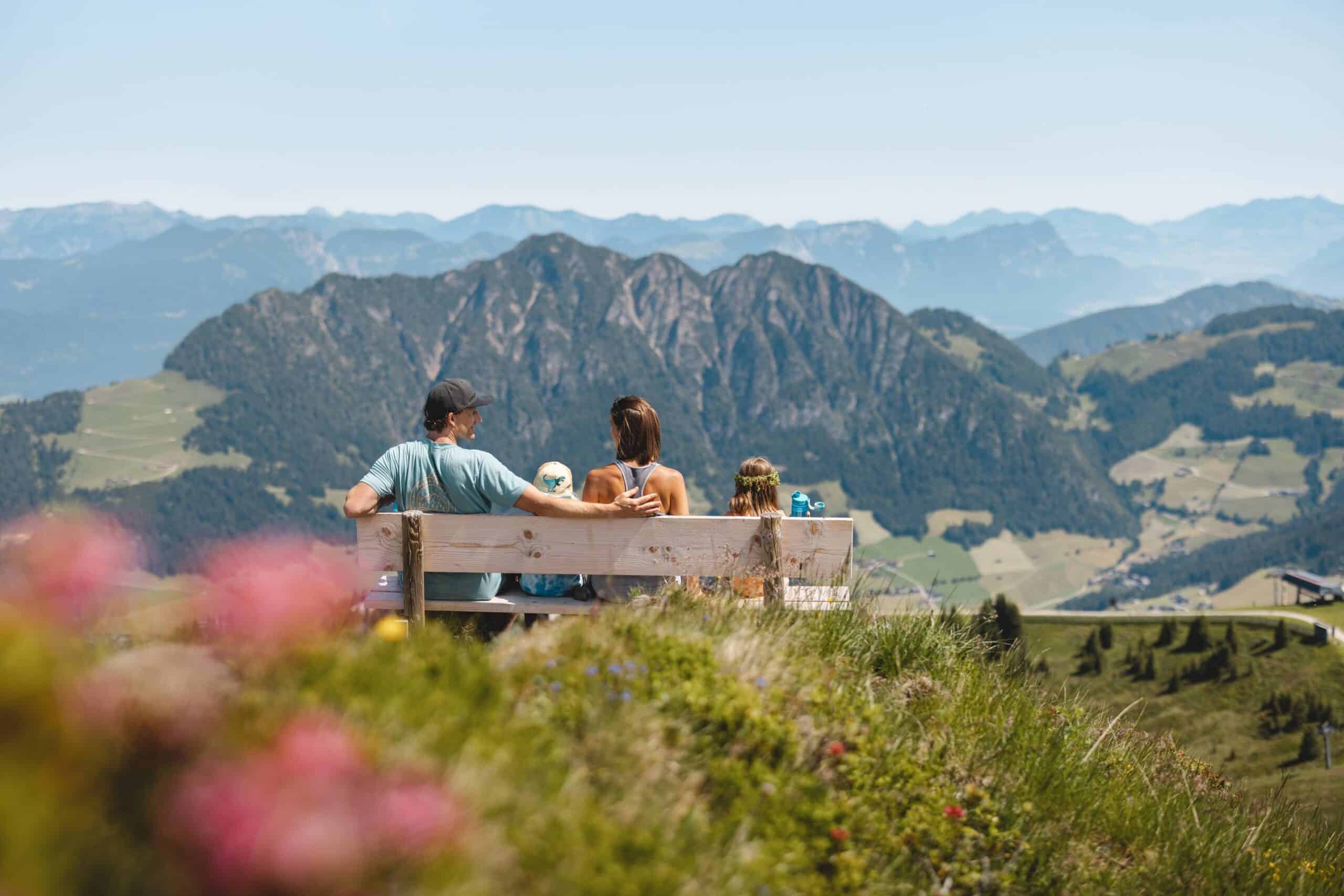 Familie betrachtet die Aussicht auf Berge von einer Holzbank aus, umgeben von grünen Feldern und blühenden Blumen an einem sonnigen Tag.