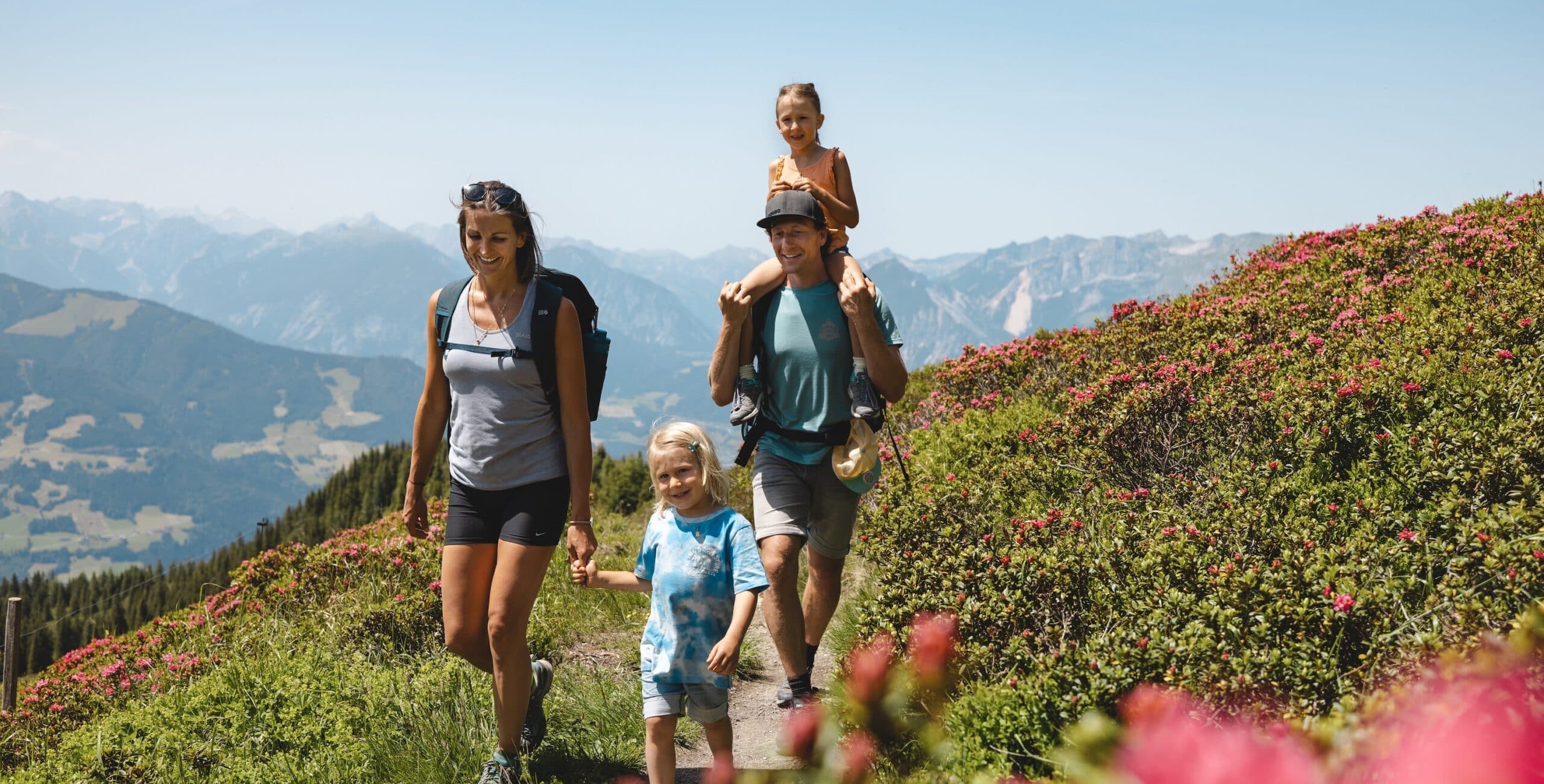 Eine Familie wandert auf einem sonnigen Pfad in den Bergen, umgeben von grüner Vegetation und mit einem Panorama von Bergketten im Hintergrund. Ein Kind wird auf den Schultern eines Erwachsenen getragen.