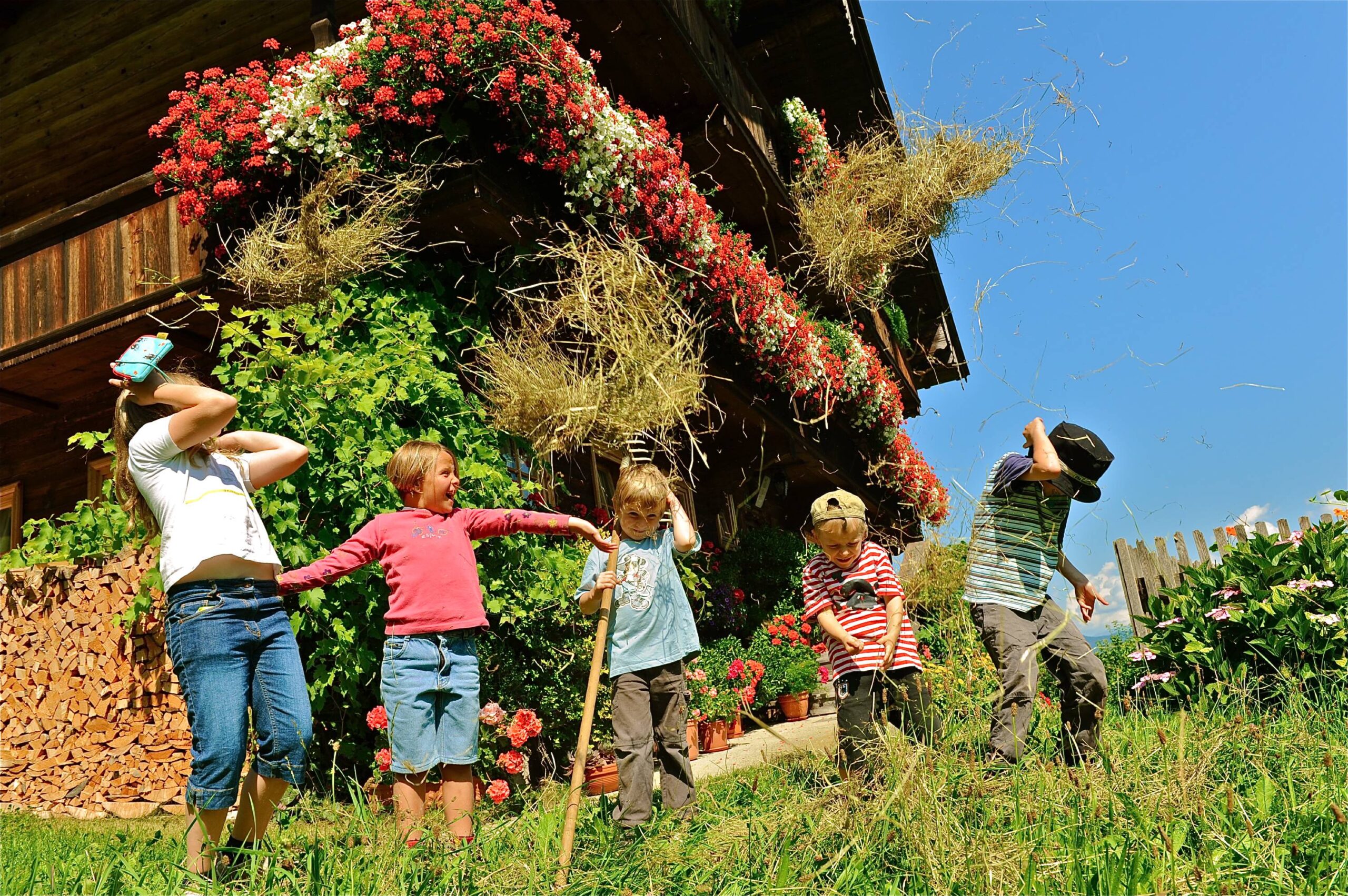 Fünf Kinder spielen vor einer Scheune auf einem Bauernhof und werfen Heu in die Luft, während im Hintergrund Blumen und grünes Gras zu sehen sind.