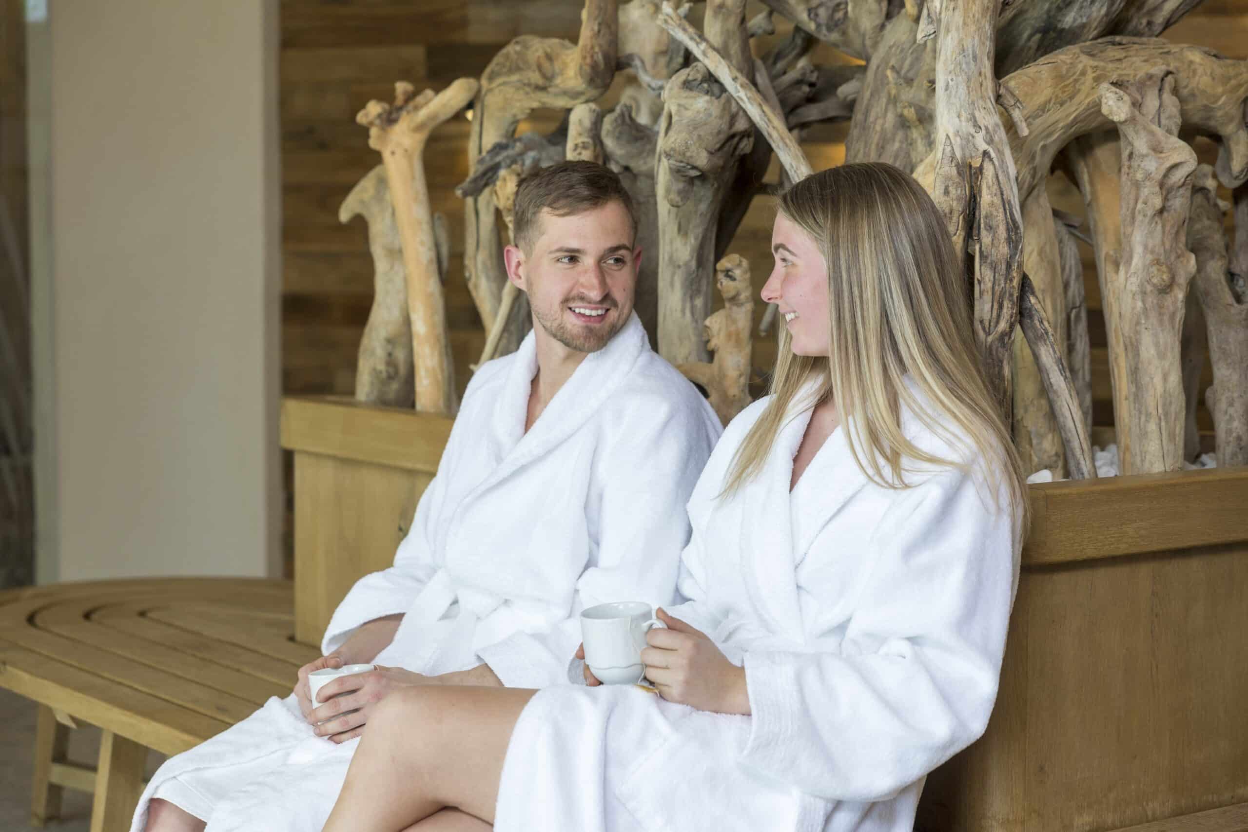 Ein Mann und eine Frau in weißen Bademänteln sitzen lächelnd auf einer Holzbank in einer Sauna, umgeben von rustikalen Holzelementen. Die Frau hält eine weiße Tasse.