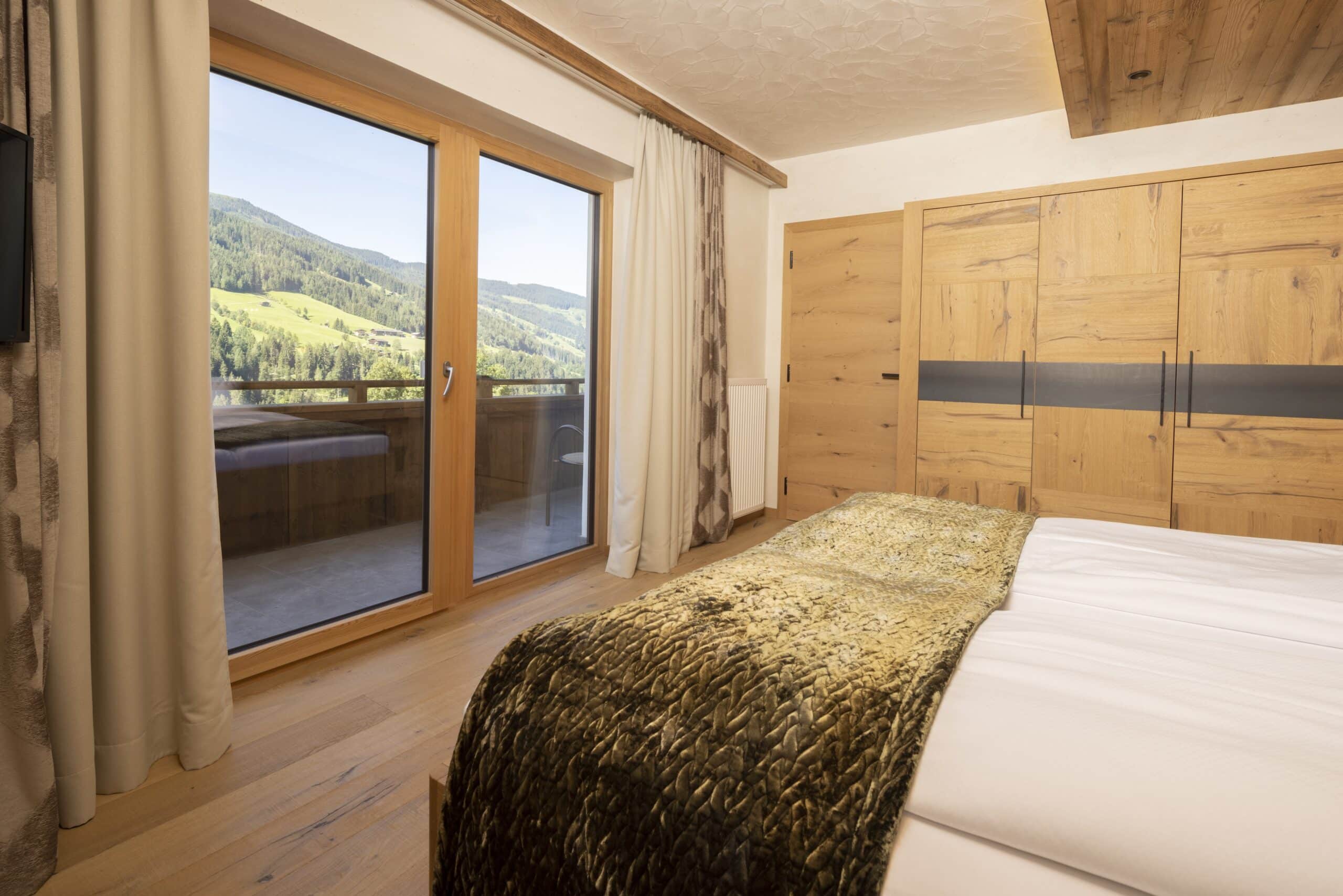 Ein gemütliches Schlafzimmer mit einem großen Bett, Holzwänden und einem großen Fenster, durch das man einen Blick auf grüne Hügel und einen blauen Himmel hat.