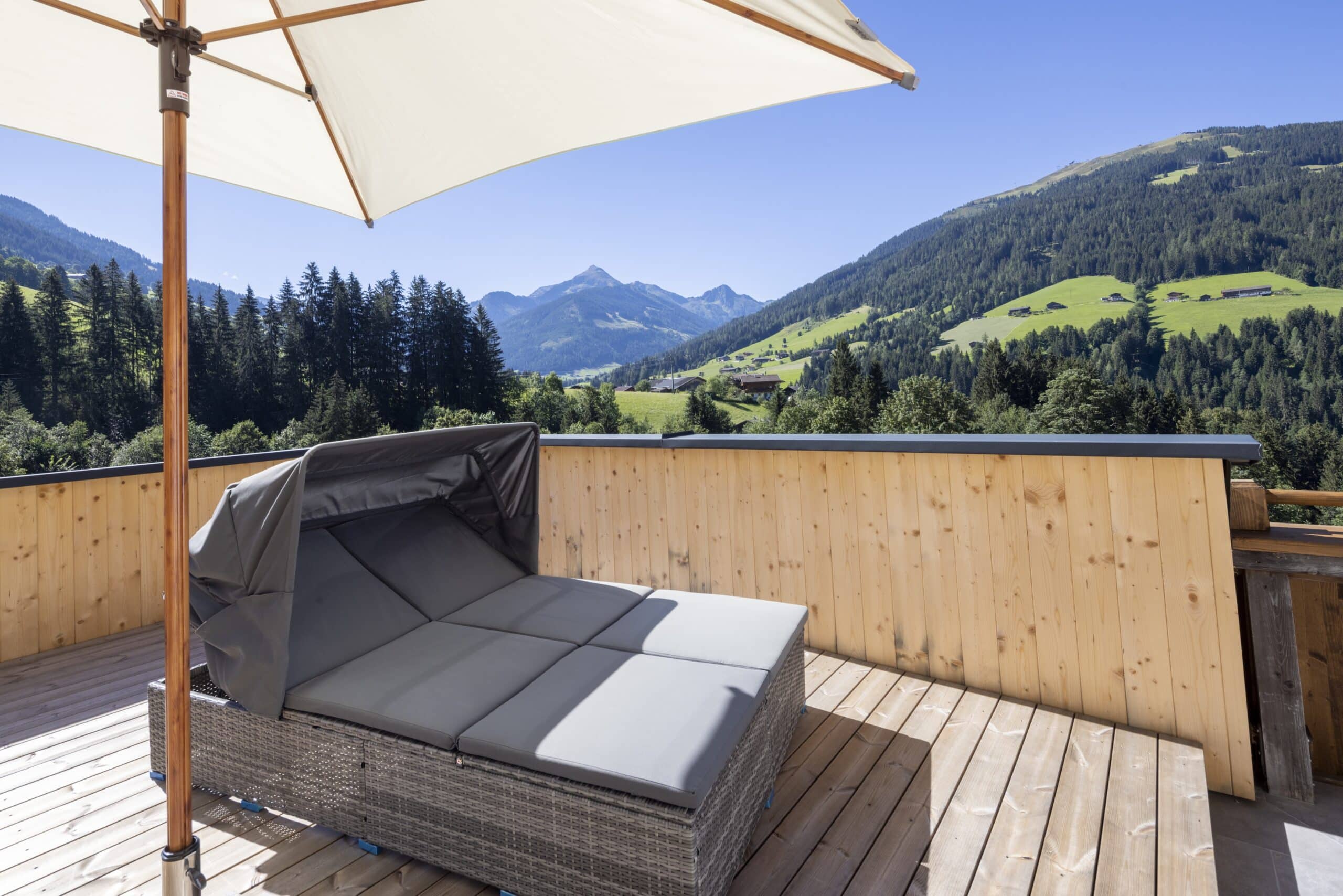 Terrasse mit Rattan-Liegesofa und Sonnenschirm vor einer malerischen Berglandschaft im Hintergrund.