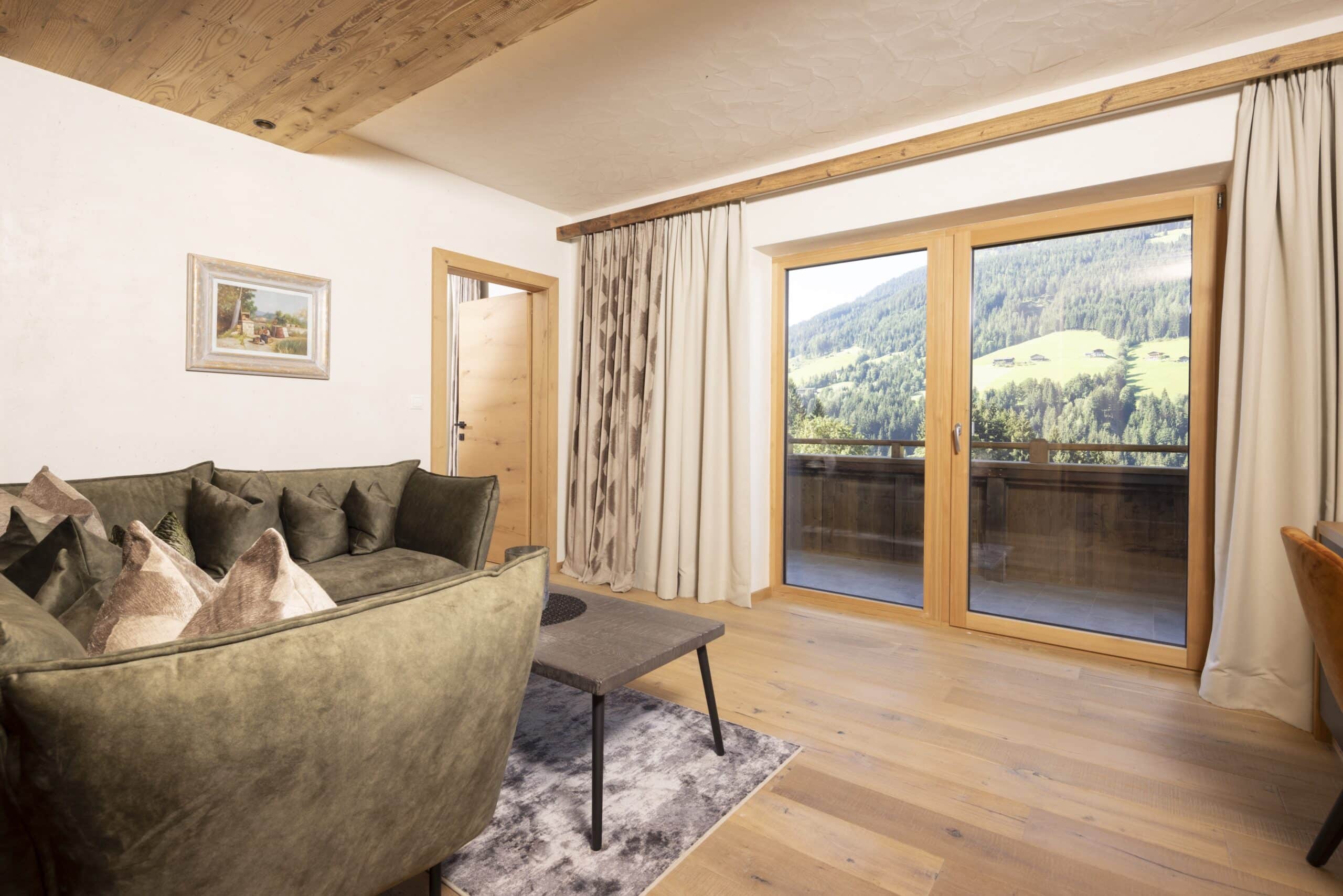 Helles Wohnzimmer mit einem großen Sofa, einem Kaffeetisch und einem flauschigen Teppich. Große Fenstertüren zeigen eine Aussicht auf grüne Hügel.