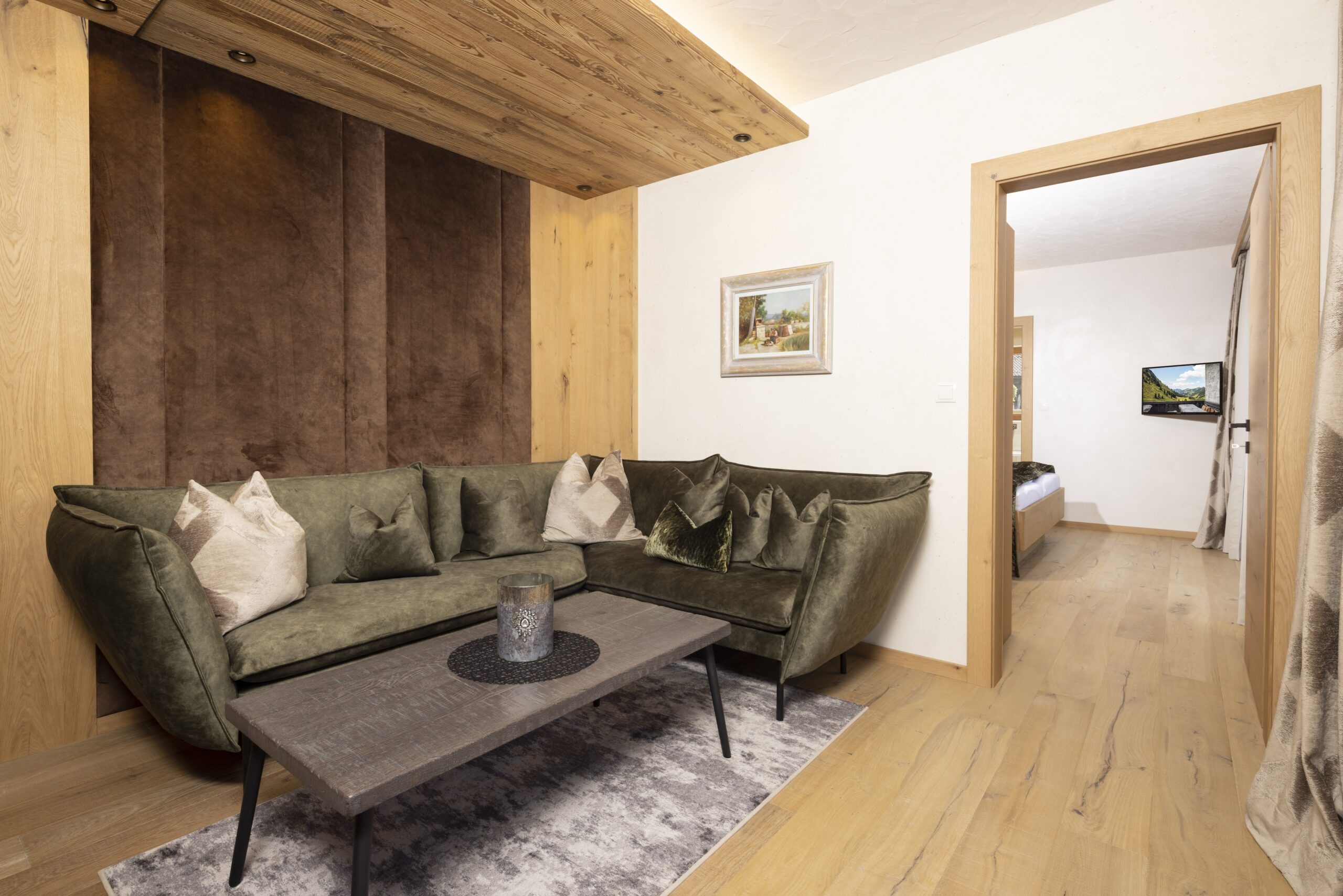 Gemütliches Wohnzimmer mit einer grünen Samtcouch, Kissen, einem Holztisch und Holzböden. Ein Fernseher ist an der Wand gegenüber der Couch angebracht.