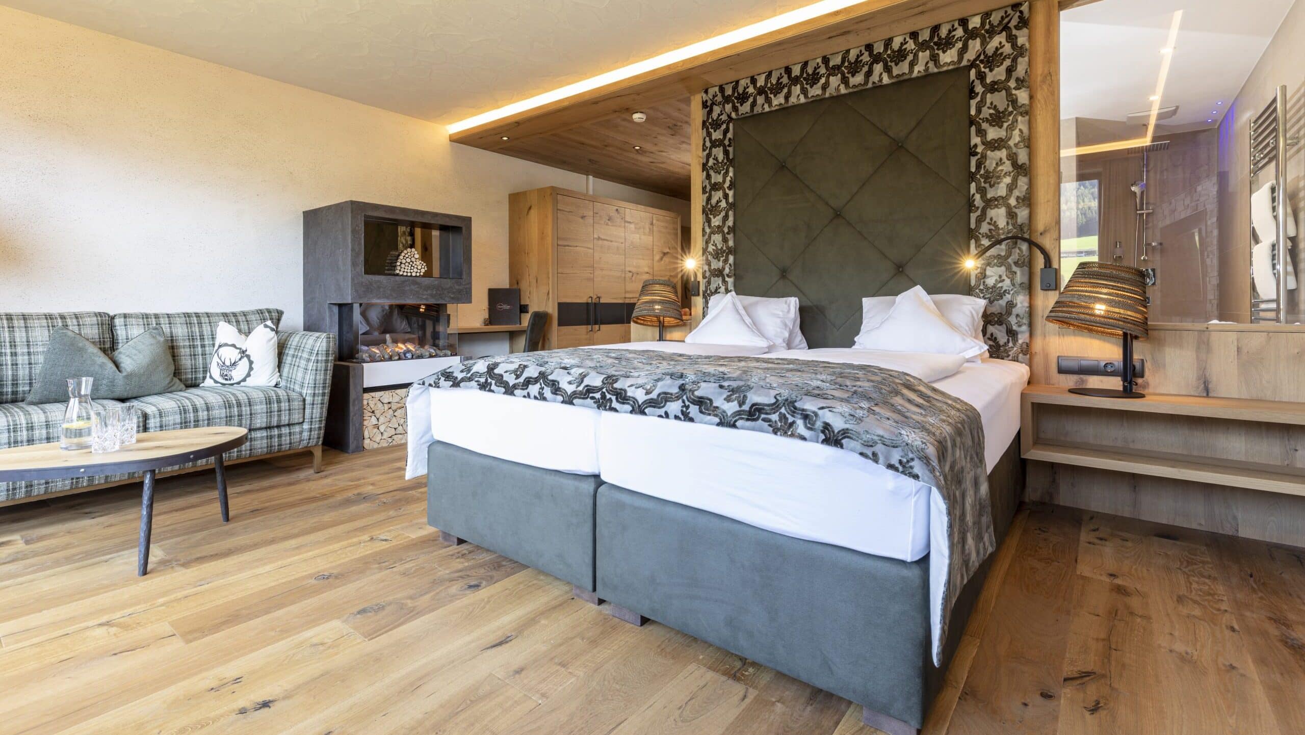 Ein stilvolles Schlafzimmer mit einem großen Doppelbett, grün gemusterter Bettwäsche und einer gemütlichen Sitzecke mit Sofa. Der Raum ist modern eingerichtet mit holzverkleideten Wänden und einem eleganten Parkettboden.