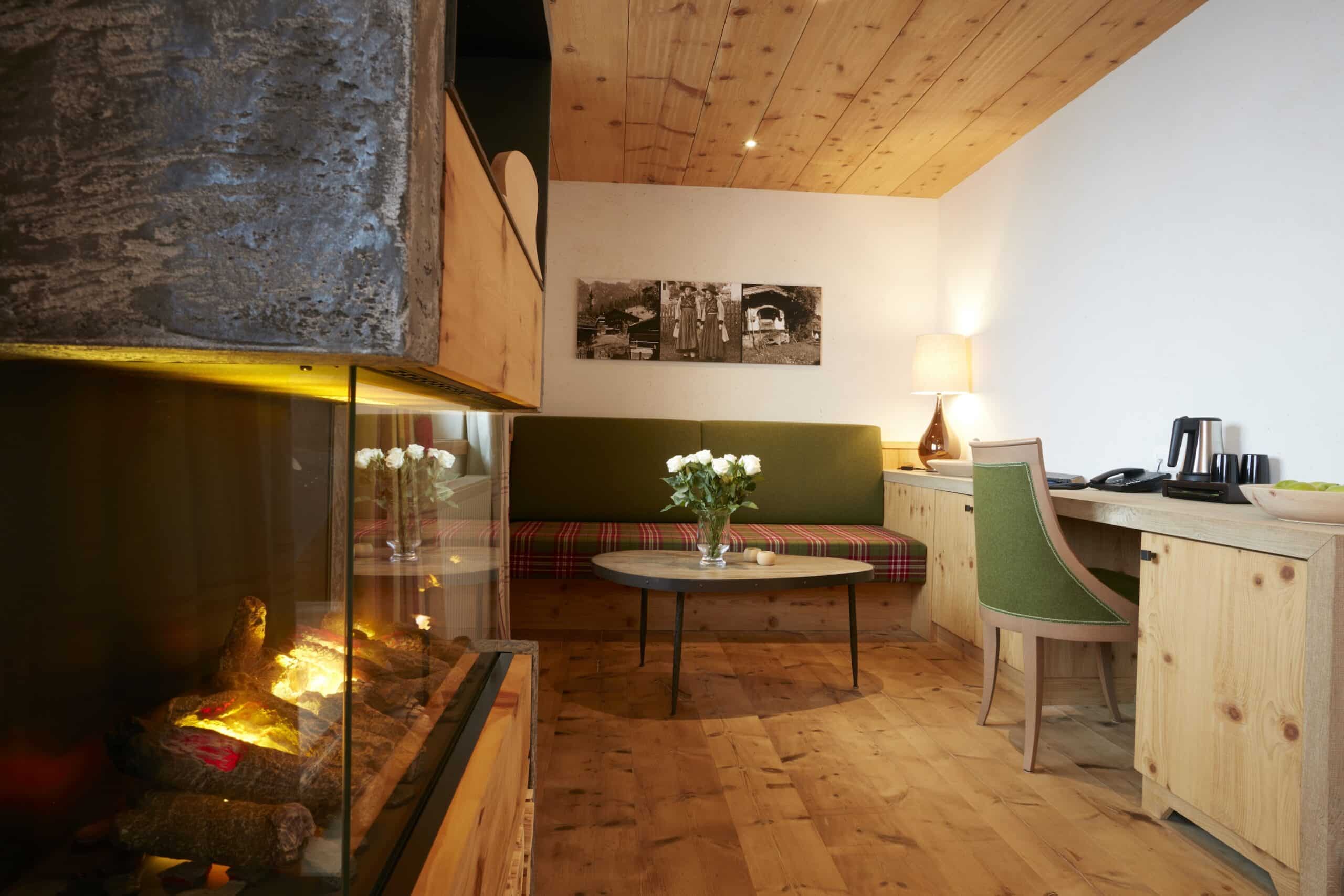 Gemütliches Wohnzimmer mit Holzverkleidung, einer Sitzecke mit grün-rotem Sofa, einem kleinen Holztisch und einer Schreibecke mit Lampe und Stuhl. Ein Kamin ist im Vordergrund sichtbar.