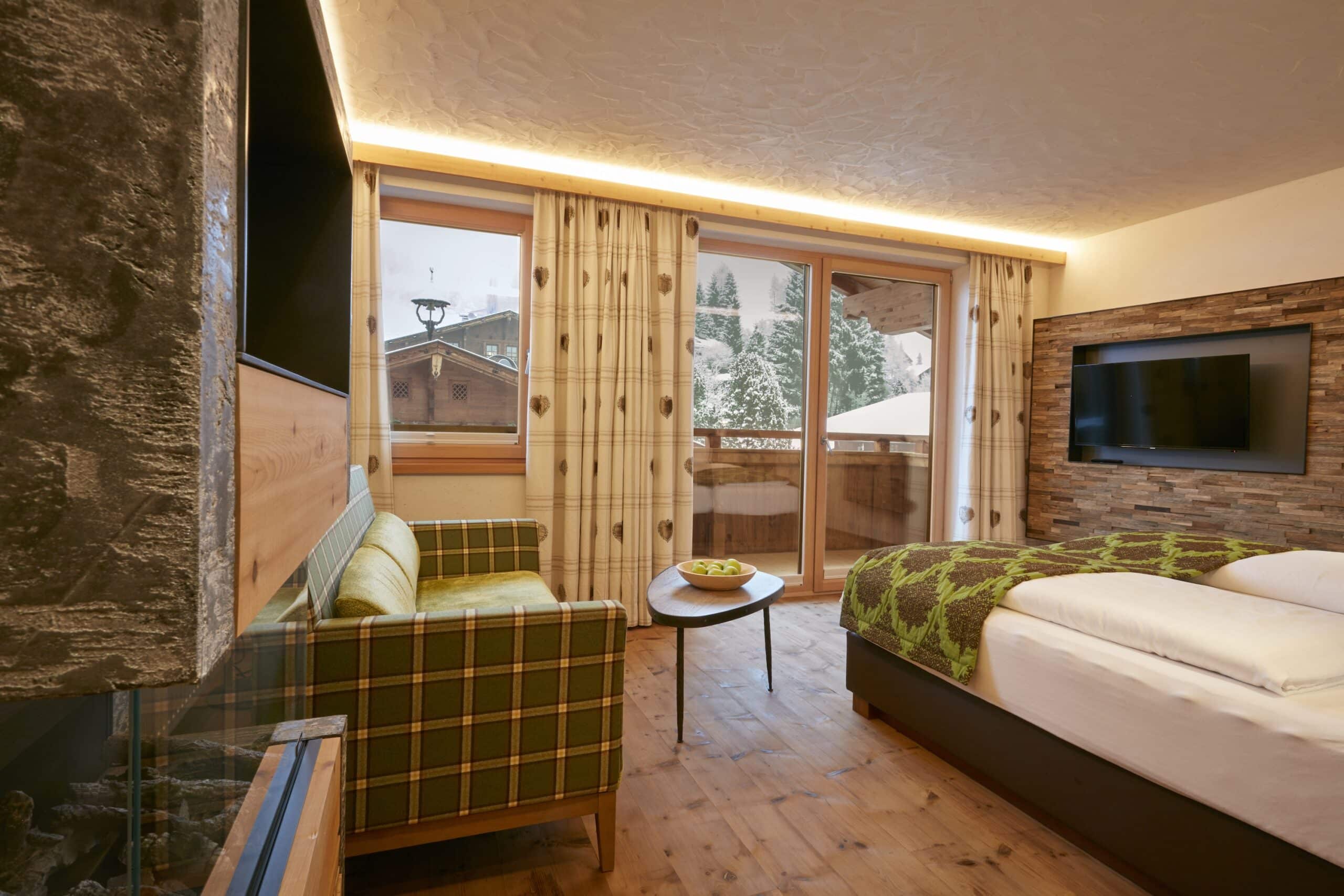 Gemütliches Hotelzimmer mit Holzfußboden und moderner Einrichtung, großes Fenster mit Blick auf verschneite Landschaft, Bett und Sofa vorhanden.