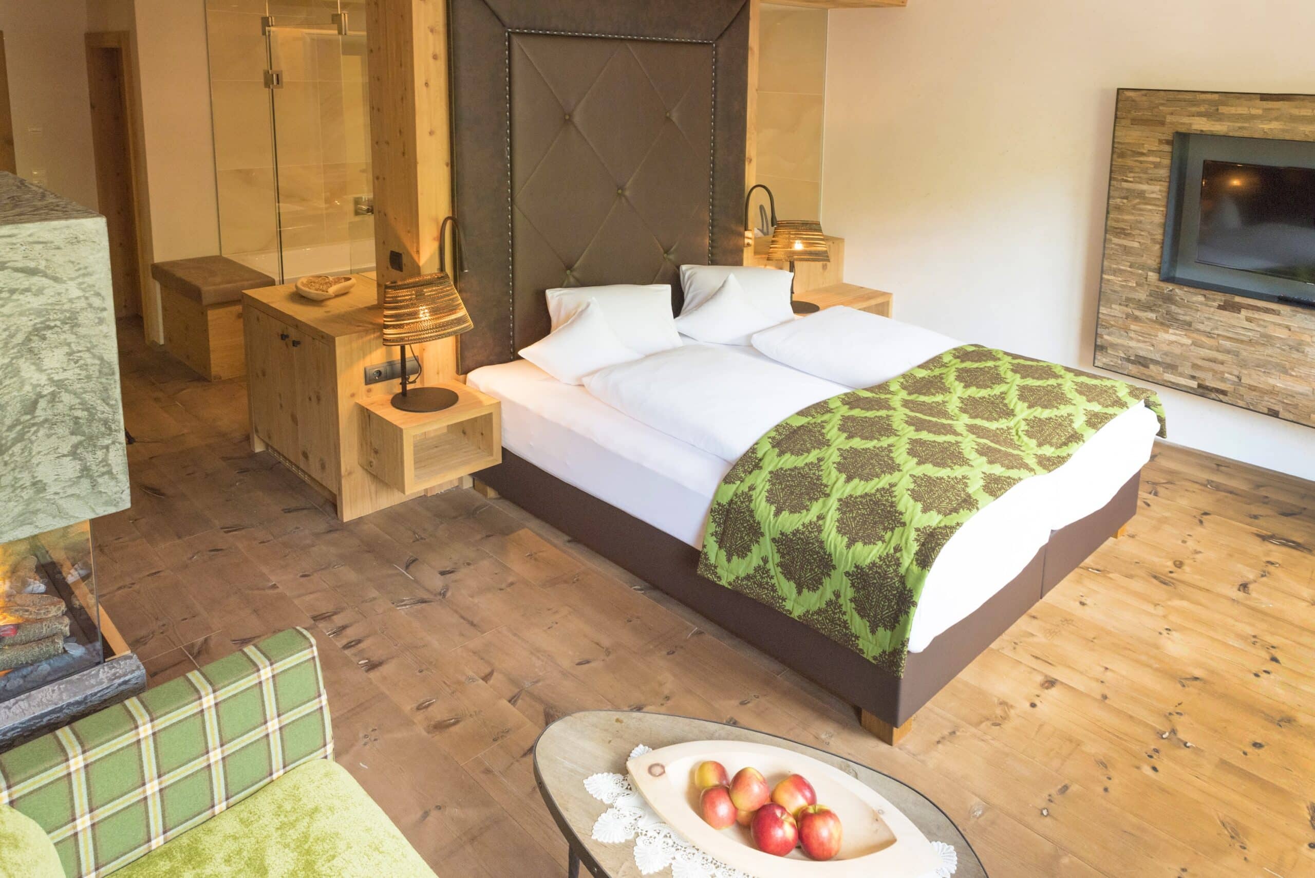 Einladendes Schlafzimmer mit einem großen Doppelbett, grün gemusterter Bettdecke und rustikaler Holzeinrichtung, einschließlich eines Holzfußbodens, einer Feuerstelle und einer modernen Saunakabine im Hintergrund.