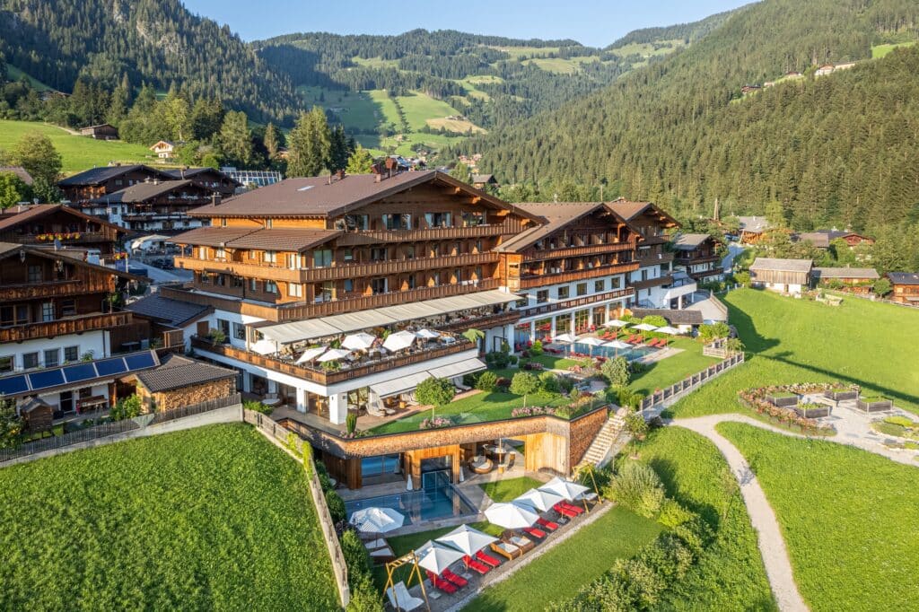 Luftaufnahme eines großen Hotelkomplexes im Chalet-Stil mit einem Außenpool, umgeben von grünen Wiesen und einer malerischen Hügellandschaft im Hintergrund.