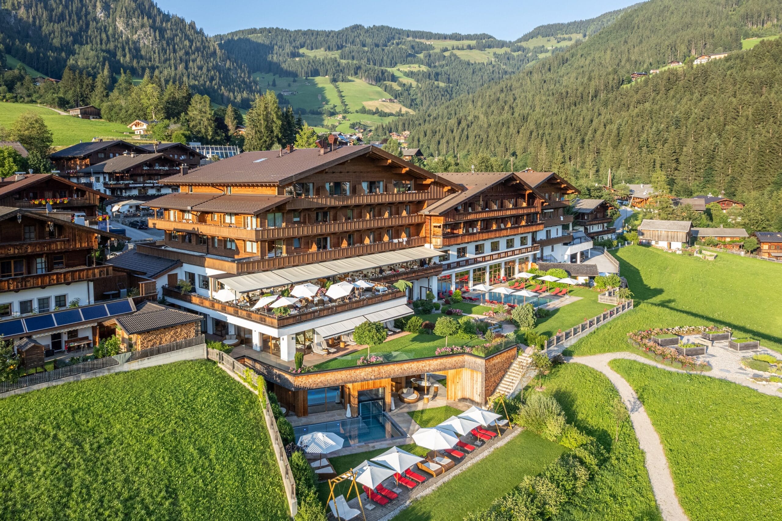 Luftaufnahme eines großen Hotelkomplexes im Chalet-Stil mit einem Außenpool, umgeben von grünen Wiesen und einer malerischen Hügellandschaft im Hintergrund.