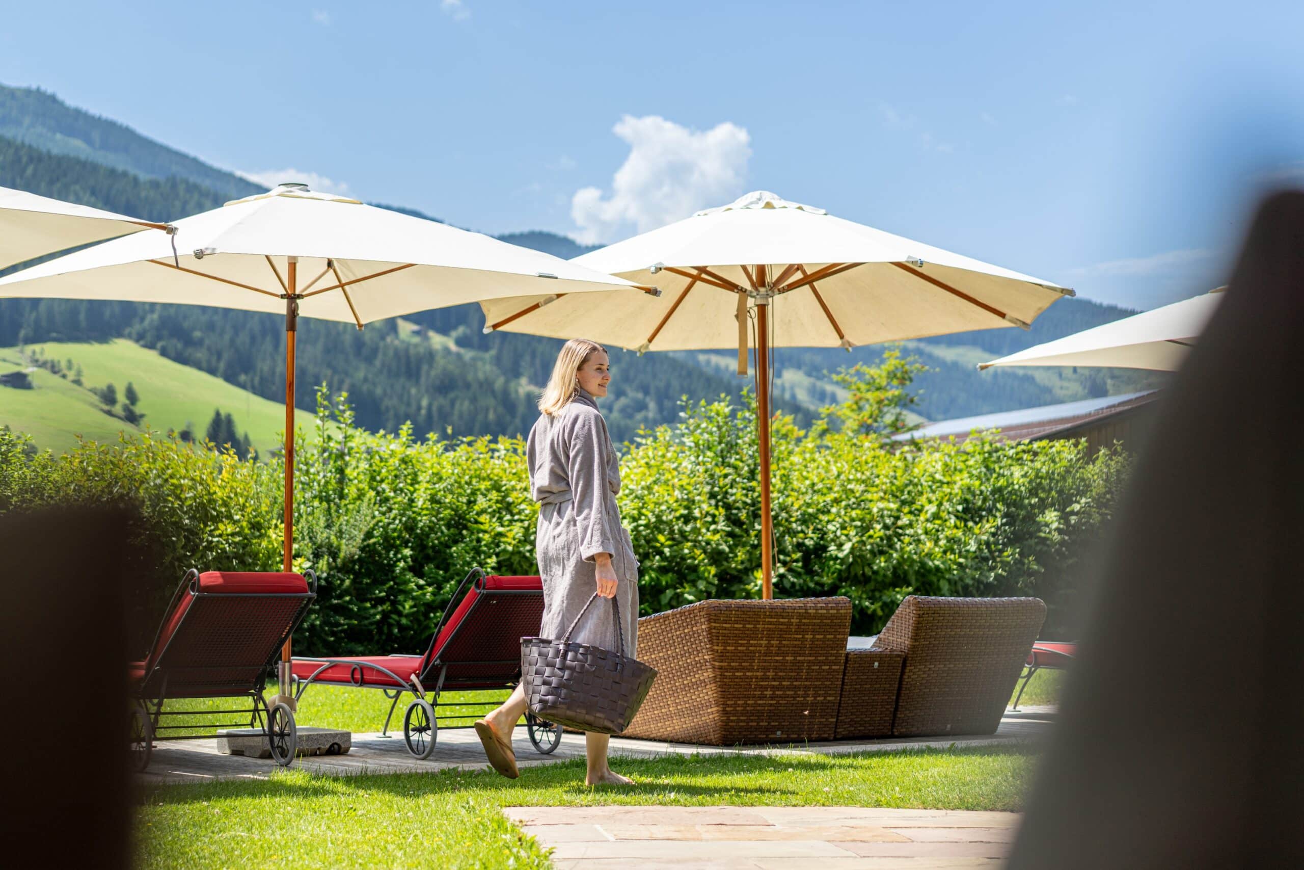 Frau im Bademantel mit Wellnesstasche im Garten - rote Liegen, beige Schirme und viel Grün, Hintergrund Berge und blauer Himmel