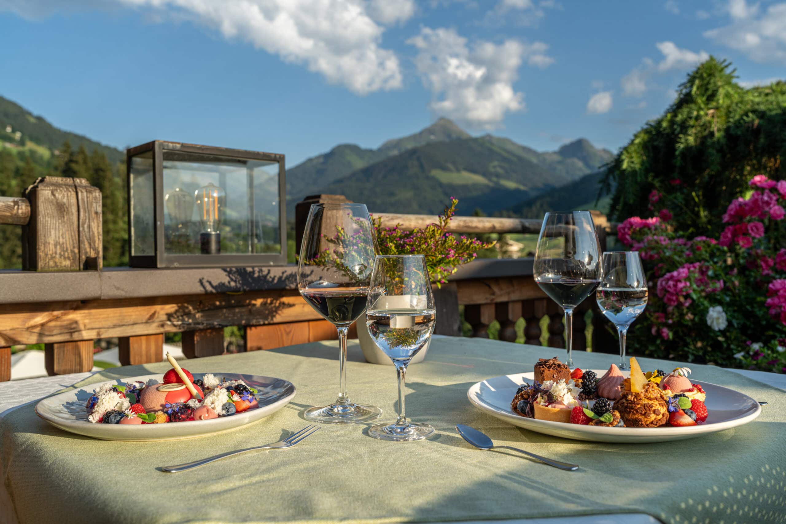 Gedeckter Tisch auf der Terrasse mit zwei angerichteten Tellern mit Desser, Rotwein- und Wasserglas, im Hintergrund Aussicht auf die Berge