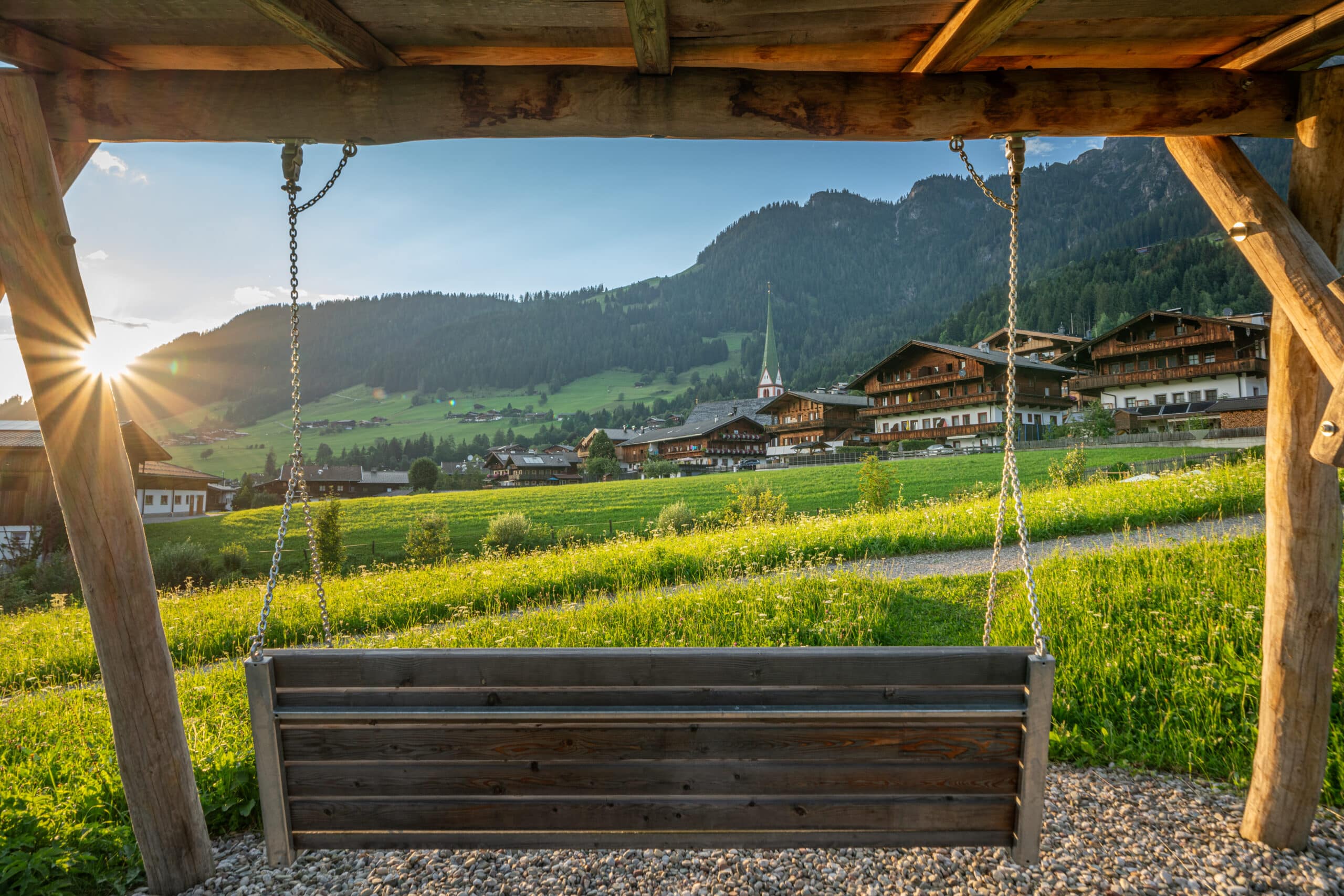 Blick auf ein idyllisches Alpendorf durch eine Holzschaukel, mit grünen Wiesen im Vordergrund und Bergen im Hintergrund bei Sonnenuntergang.