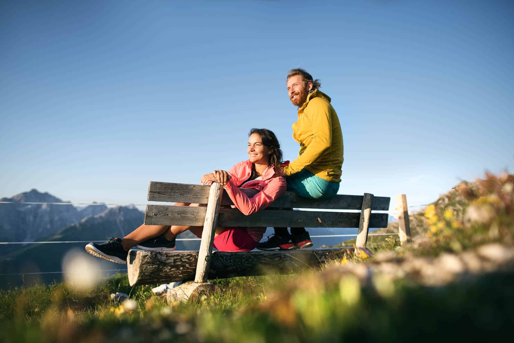 Pärchen sitzt auf einer Bank, Sonne im Gesicht, am Berg, Hintergrund Berge, blauer Himmel