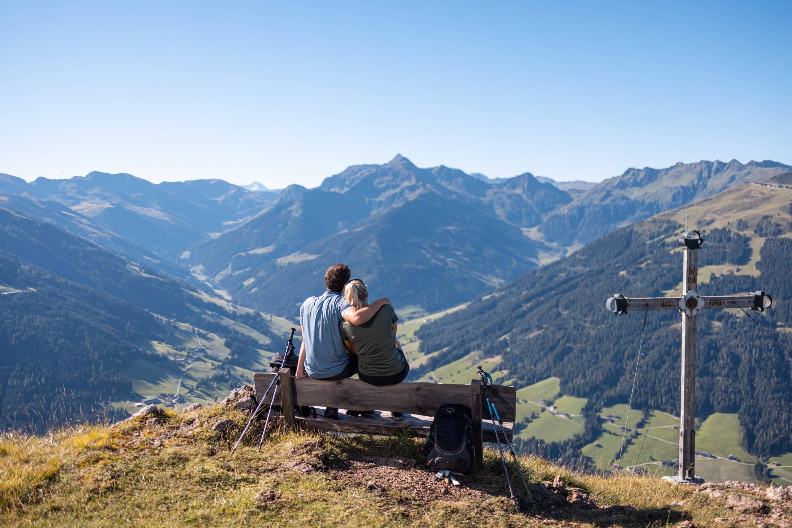 Pärchen sitzt aneinandergekuschelt auf einer Bank auf dem Berggipfel. Daneben ein kleines Bergkreuz. Ausblick auf das ganze Tal und die Berwelt