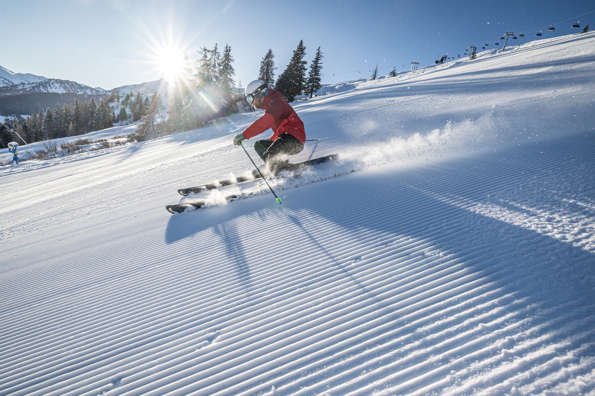 Skifahrer in roter Skijacke auf frisch präparierter Piste, blauer Himmer, Sonne blitzt durch
