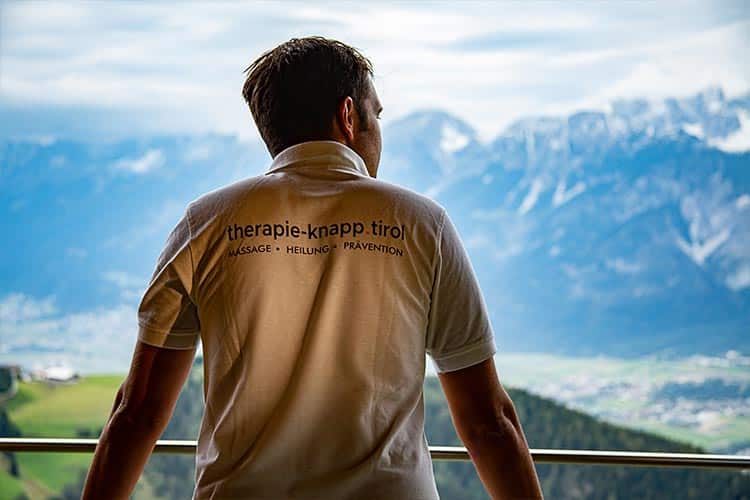 Therapeut_Johannes Knapp-mit weißem Shirt und Logoaufdruck_von hinten_blickt in die Berge